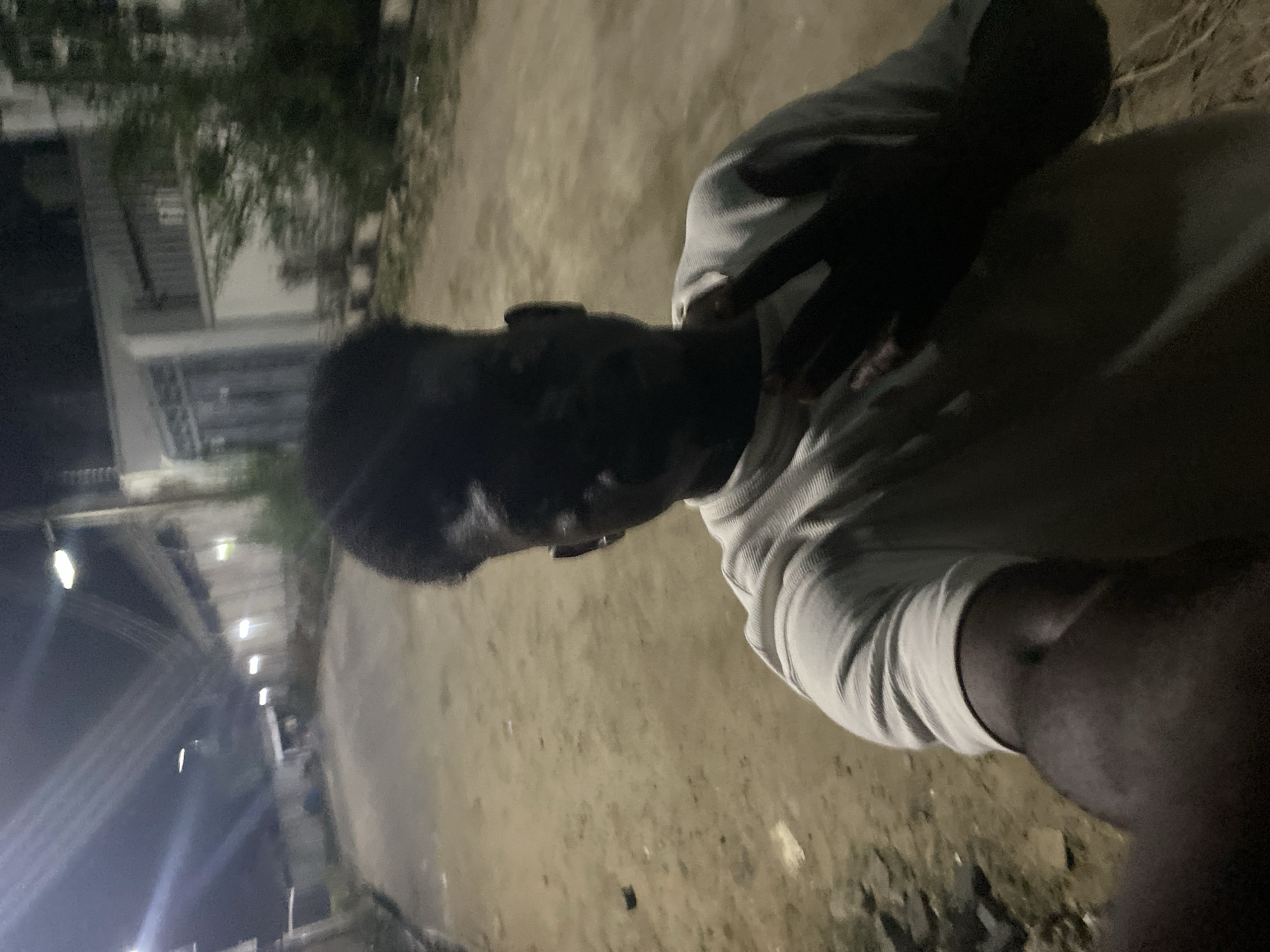 A man stands in an outdoor, urban environment at night. The man, who has dark skin, wears a light-colored, long-sleeved shirt. His right hand is raised near his neck. The scene is dimly lit by distant street lights. In the background are buildings and greenery. The location is in the city of Unknown, Unknown.