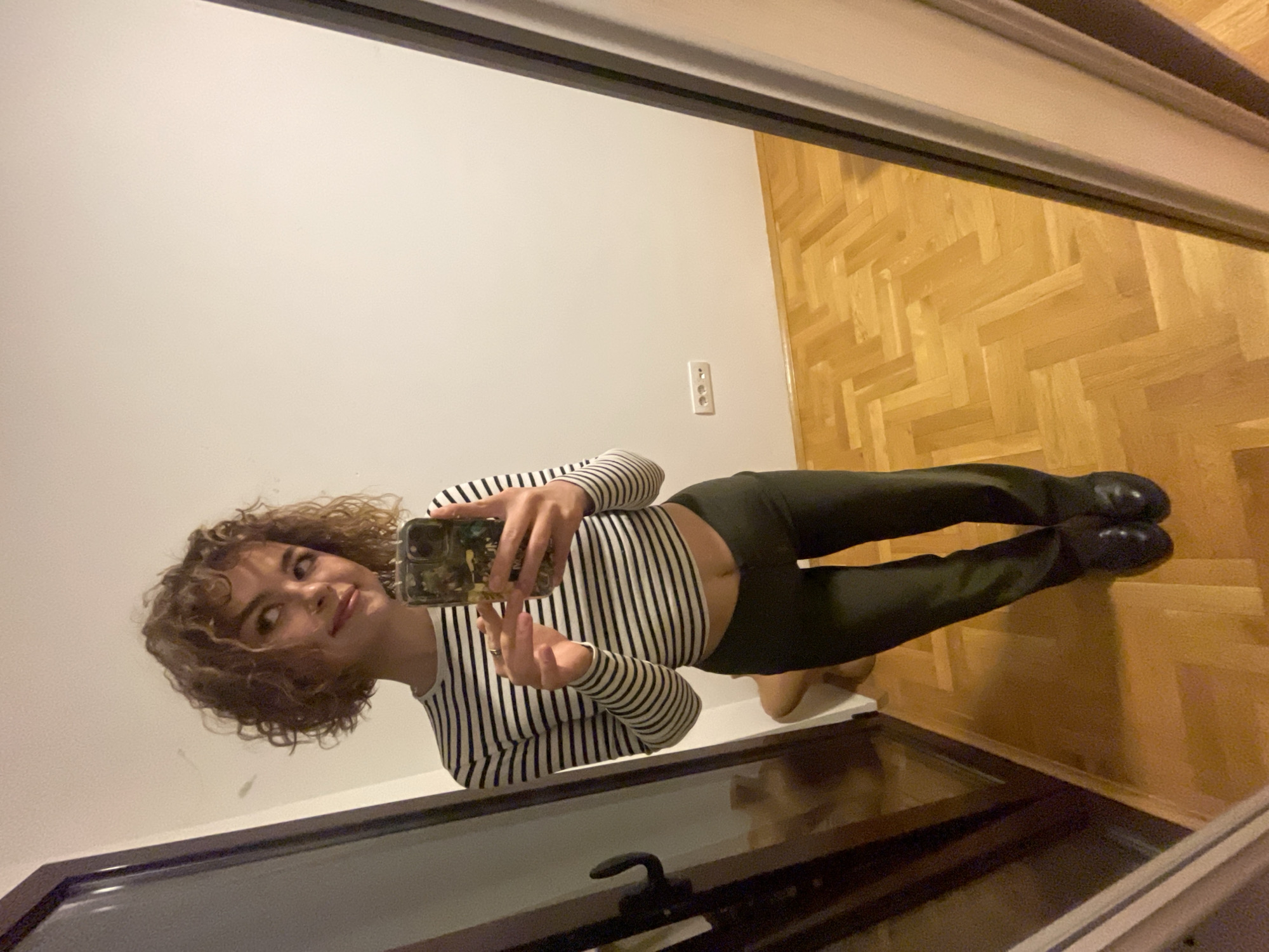 A young woman is taking a selfie in front of a mirror. She is wearing a striped long-sleeved shirt and dark pants, and black shoes. She has curly hair. The room has light wood flooring. The wall is white with a light switch. The location is Boreti, Montenegro.