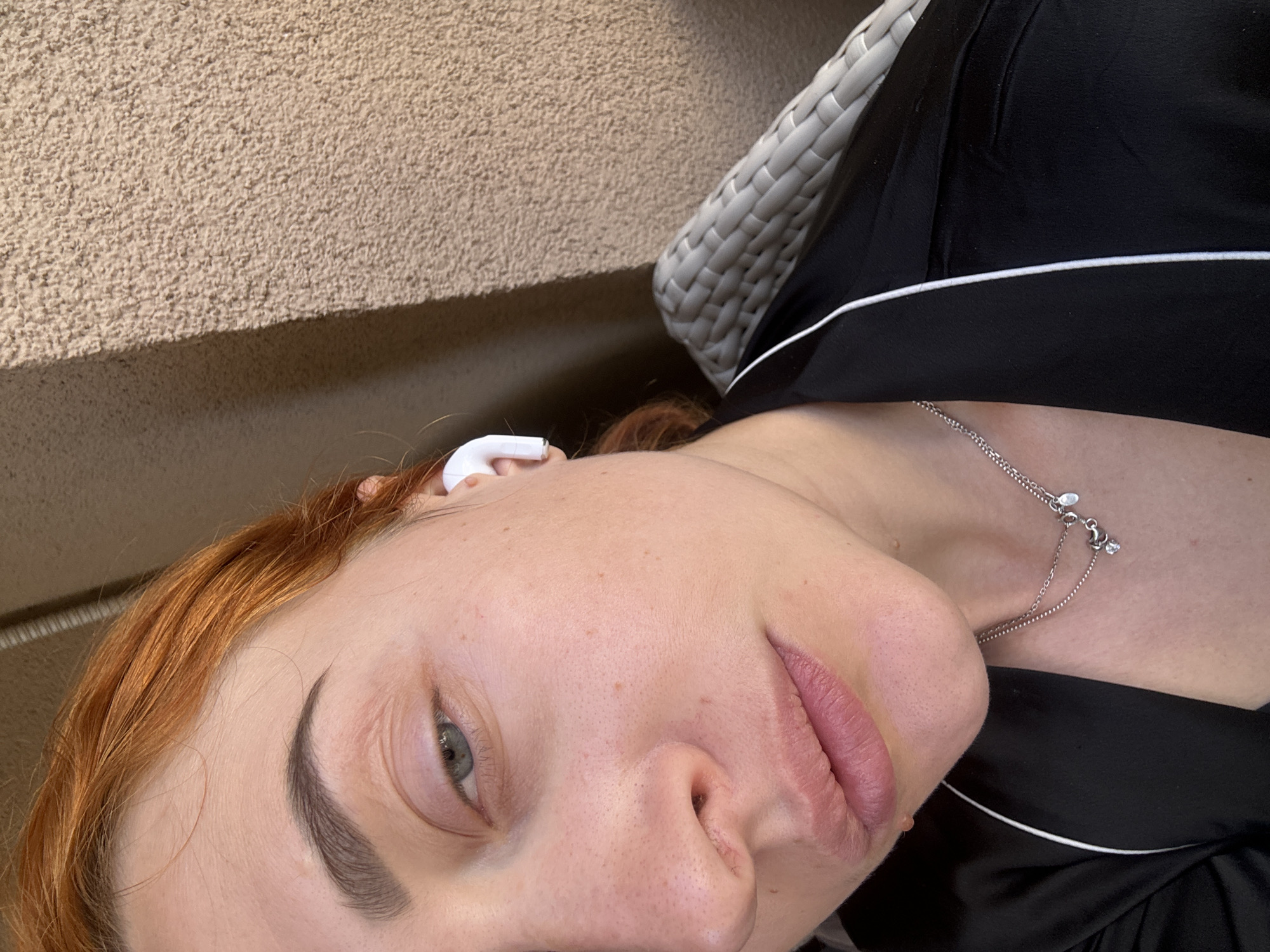 A close-up, angled shot shows a fair-skinned woman with red hair and an AirPod in her ear. She has grey eyes and is wearing a silver necklace. She is wearing black pajamas with white trim. The background shows a beige textured wall and a white woven chair. The location is potentially Budva, Montenegro.