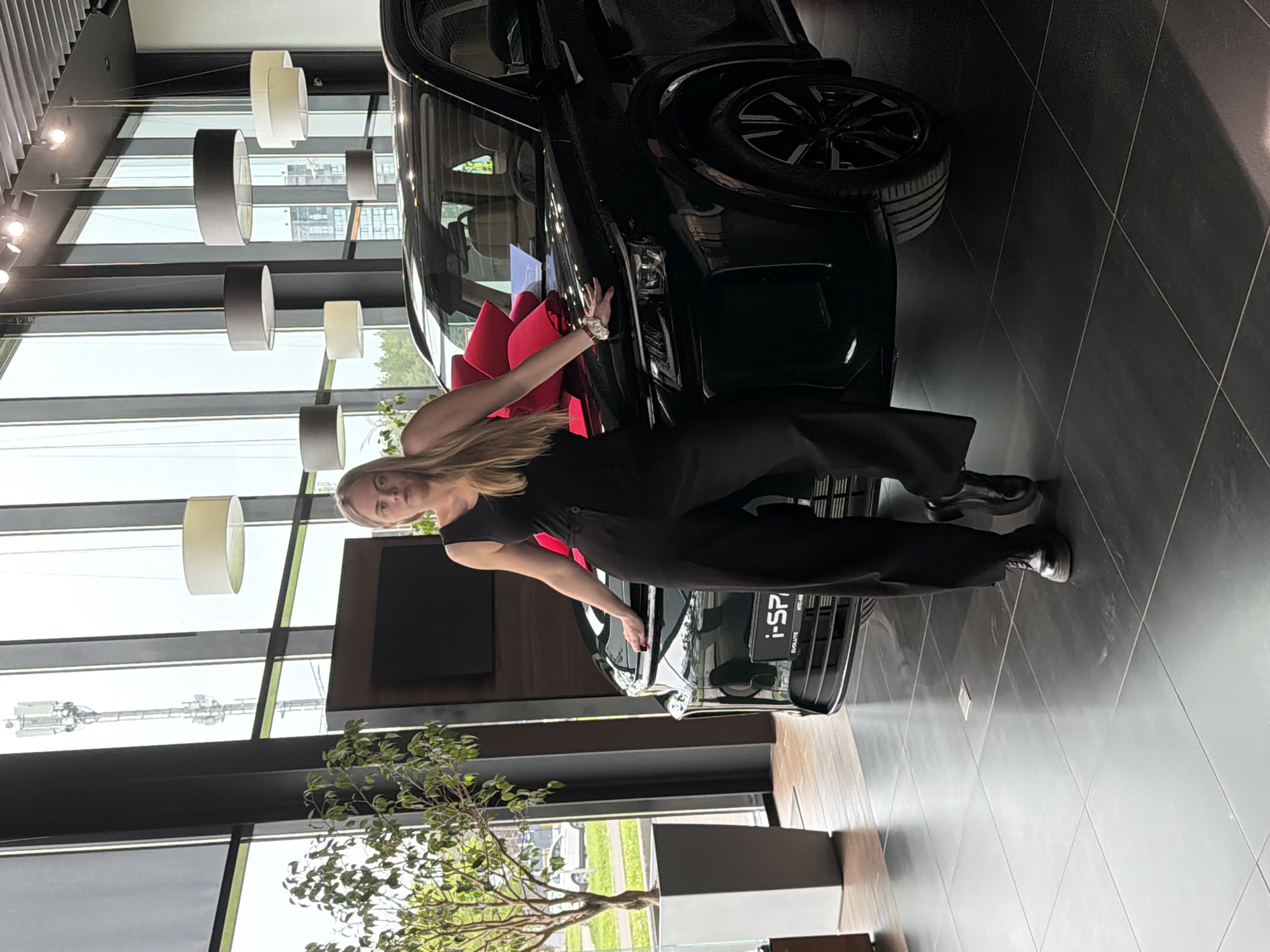 The image shows a woman standing next to a black car in what appears to be a car dealership. The woman is wearing a black jumpsuit and black shoes. The car has a large red bow on the hood. A potted tree is in the foreground. Through the windows, a city landscape with buildings and a cell tower is visible. Based on textual markings on the car, the location is likely Izhevsk, Russia.