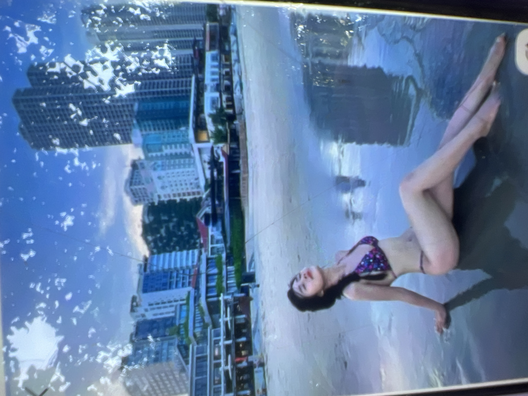 A young woman in a bikini sits on a wet beach with buildings in the background. The woman has dark hair styled in a braid and is wearing a patterned bikini. She is leaning back on her arms, with her legs extended. The beach is wet and reflective, with a calm sea. In the background, there are several modern buildings, including high-rise towers. The sky is blue with some clouds. The image appears to be taken in a city setting with a beach. The location of Kara-Dıykan, Kyrgyzstan is not visible in the image.