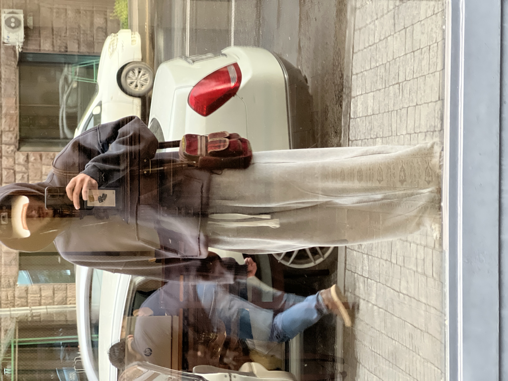 A person wearing a beige hat and a dark jacket is holding a phone and taking a photo, reflected in a window. The window also reflects a street scene in Yerevan, Armenia. A white car is parked next to the sidewalk, and the reflection shows another person inside the car with a laptop. The reflection includes the exterior of the building across the street and part of the sidewalk. The outside light suggests a cloudy day.