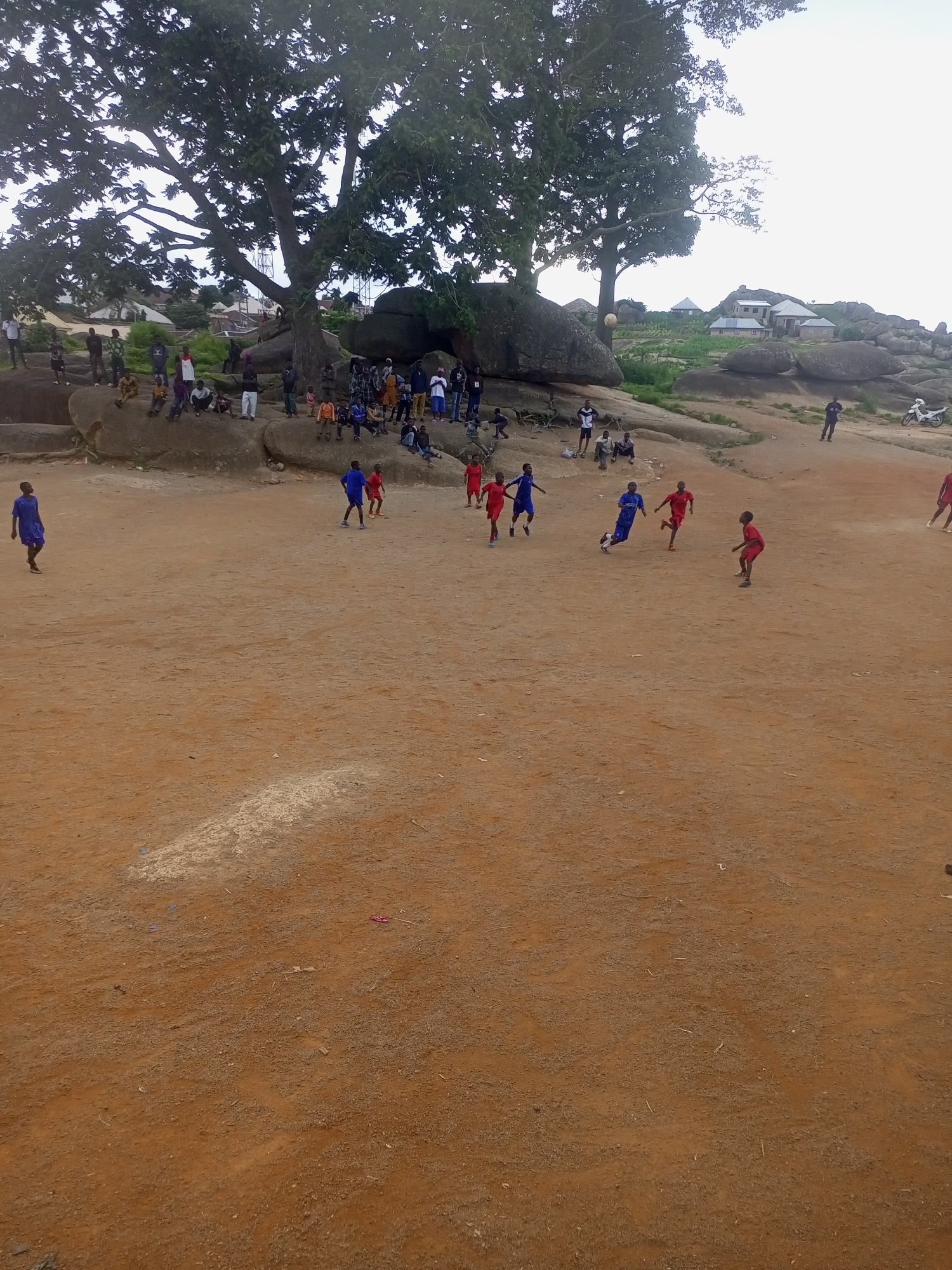 The image depicts a soccer game being played on a dirt field in Jos, Nigeria. A group of young boys in red and blue jerseys are actively engaged in the match, with several spectators gathered on a rocky incline in the background. Large trees and rocks characterize the landscape, with some buildings visible further up the hill. The overall scene suggests a community gathering for a local sporting event.