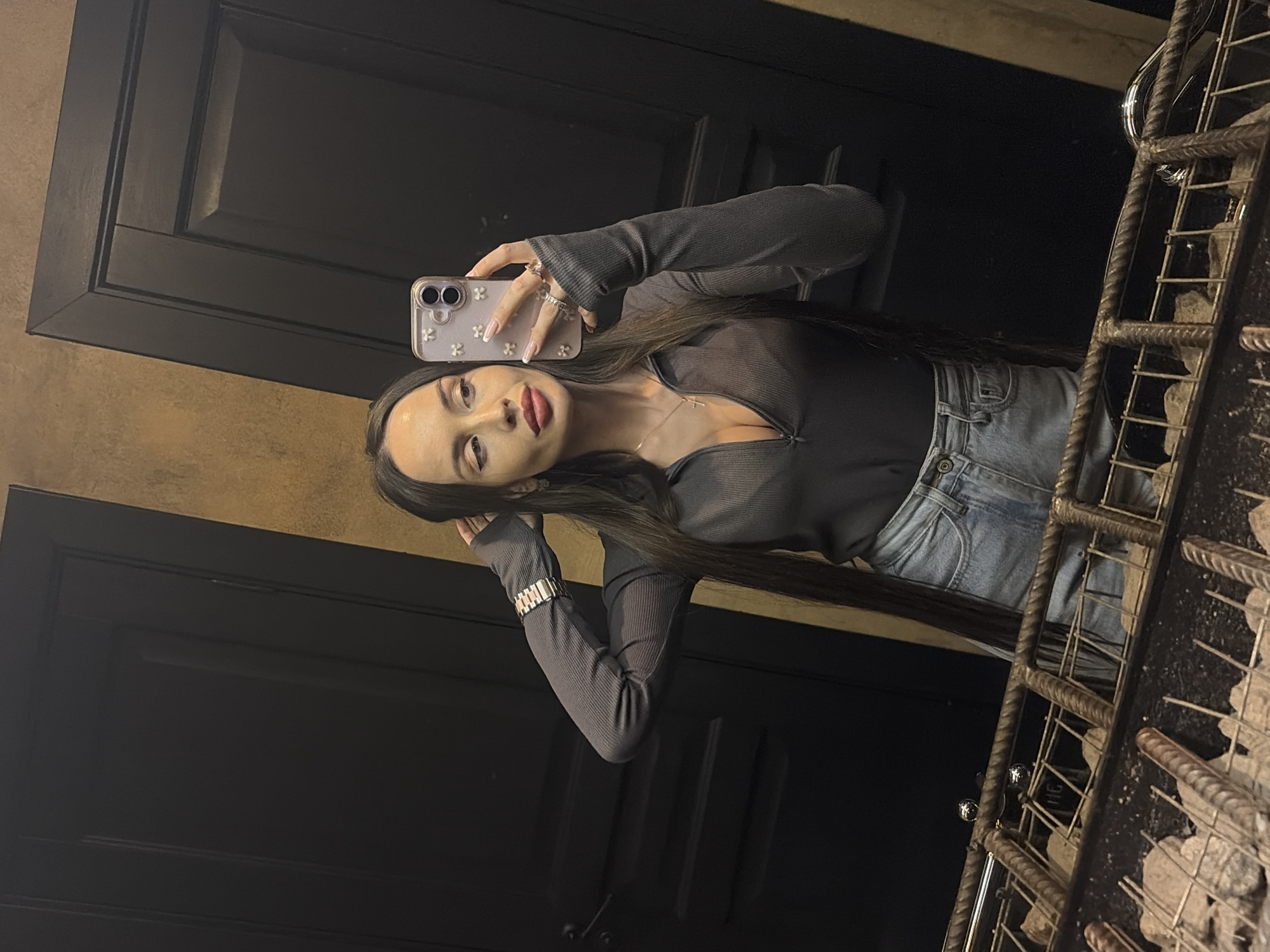 A woman takes a mirror selfie in front of a dark door in an interior setting. She wears a gray long-sleeved shirt, light blue jeans, a gold necklace, and a gold watch. She holds an iPhone with a white floral case in her left hand. The background includes a dark-colored door and a metallic grid-like structure filled with rocks, possibly a design element. Based on available information, it is not possible to determine if the location is in Saint Petersburg, Russia.