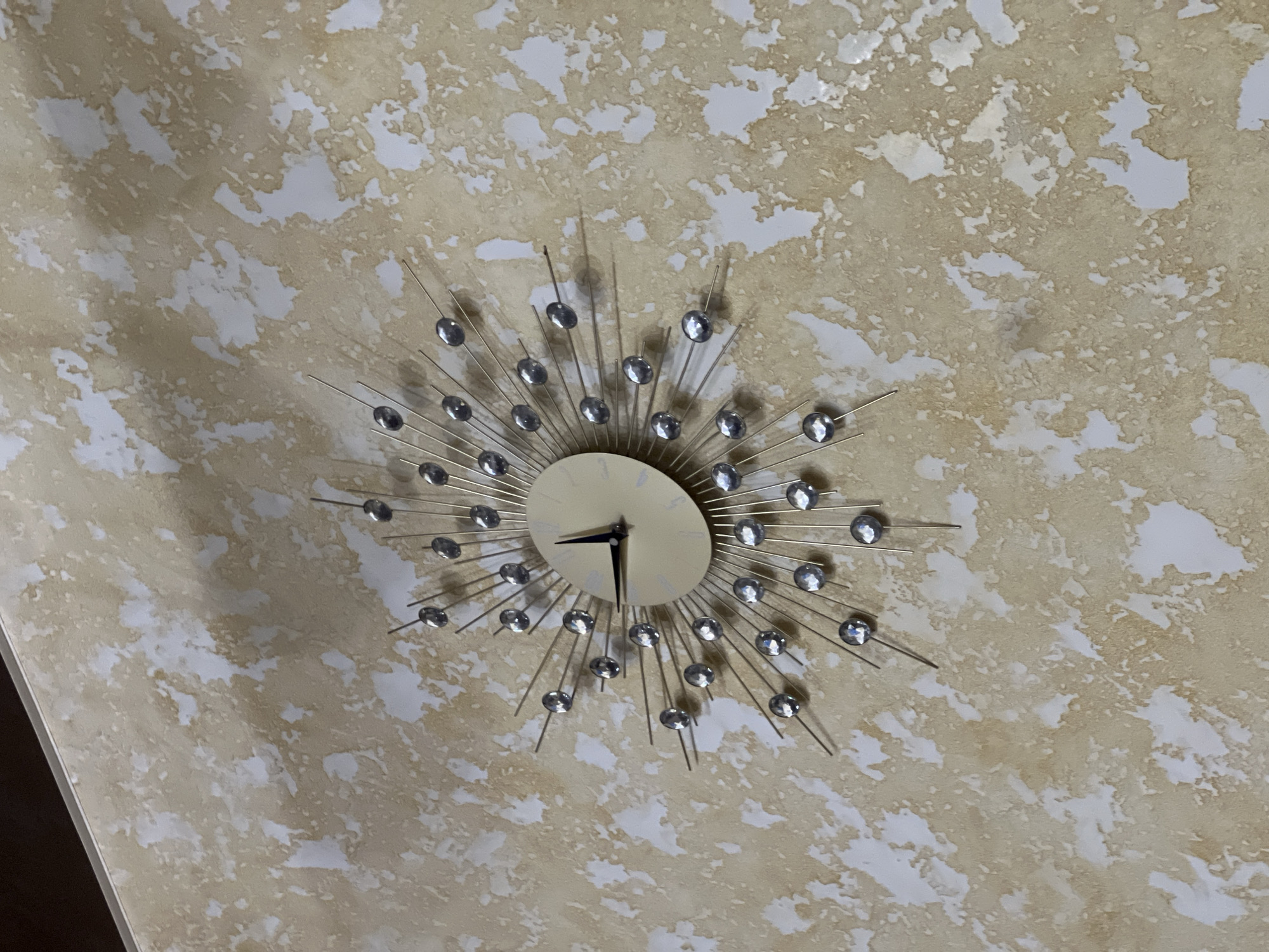 The image shows a close-up, angled shot of a decorative wall clock with a sunburst design. The clock face is circular and cream-colored, with black hour and minute hands. The clock is adorned with numerous thin metal spokes radiating outwards, each tipped with a faceted, clear embellishment that reflects light. The wall behind the clock has a textured finish with a pattern of irregular splotches in shades of beige and white. The overall impression is of an indoor domestic setting. There is no information in the image to suggest a specific location in Lviv, Ukraine.