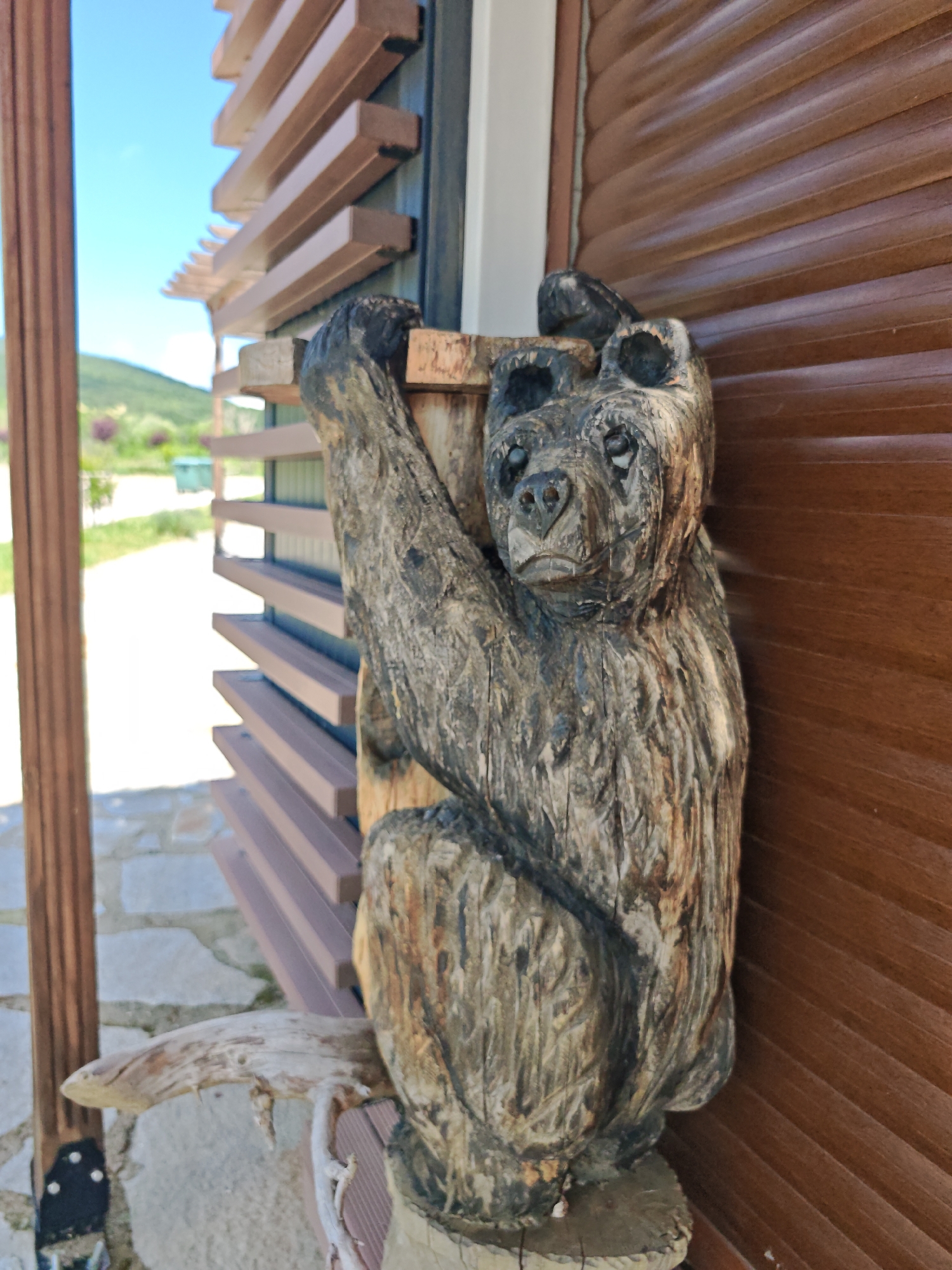 This image, captured in Svirachi, Bulgaria, displays a detailed, weathered wooden bear sculpture positioned prominently in the mid-right foreground. The bear, carved from dark brown wood with visible grain and texture replicating fur, is depicted in a clinging posture, gripping a light brown horizontal wooden plank with both front paws. Its face, with dark, hollowed eye sockets and a textured snout, is directed towards the viewer, conveying a somber expression.
To the bear's right, a vertical surface composed of numerous horizontal, overlapping dark brown wooden slats forms a background element. Behind and to the left of the bear, a section of a structure with horizontally arranged, stacked light brown wooden beams creates a slatted screen. A sturdy, vertical dark brown wooden post is situated on the far left, featuring a dark metal bracket with visible white fasteners at its base. Beneath the wooden structure and extending into the foreground, a pathway made of irregularly shaped, light gray flagstones is visible. A gnarled, light-colored piece of driftwood protrudes horizontally from the lower left of the structure's base.
The background, rendered out of focus, shows a clear, bright blue sky occupying the upper left quadrant, extending over distant green hills. Below the hills, green vegetation is interspersed with clusters of blurry purple flowering plants. A indistinct, light green object is discernible in the distant mid-ground. No human figures are present in the scene.