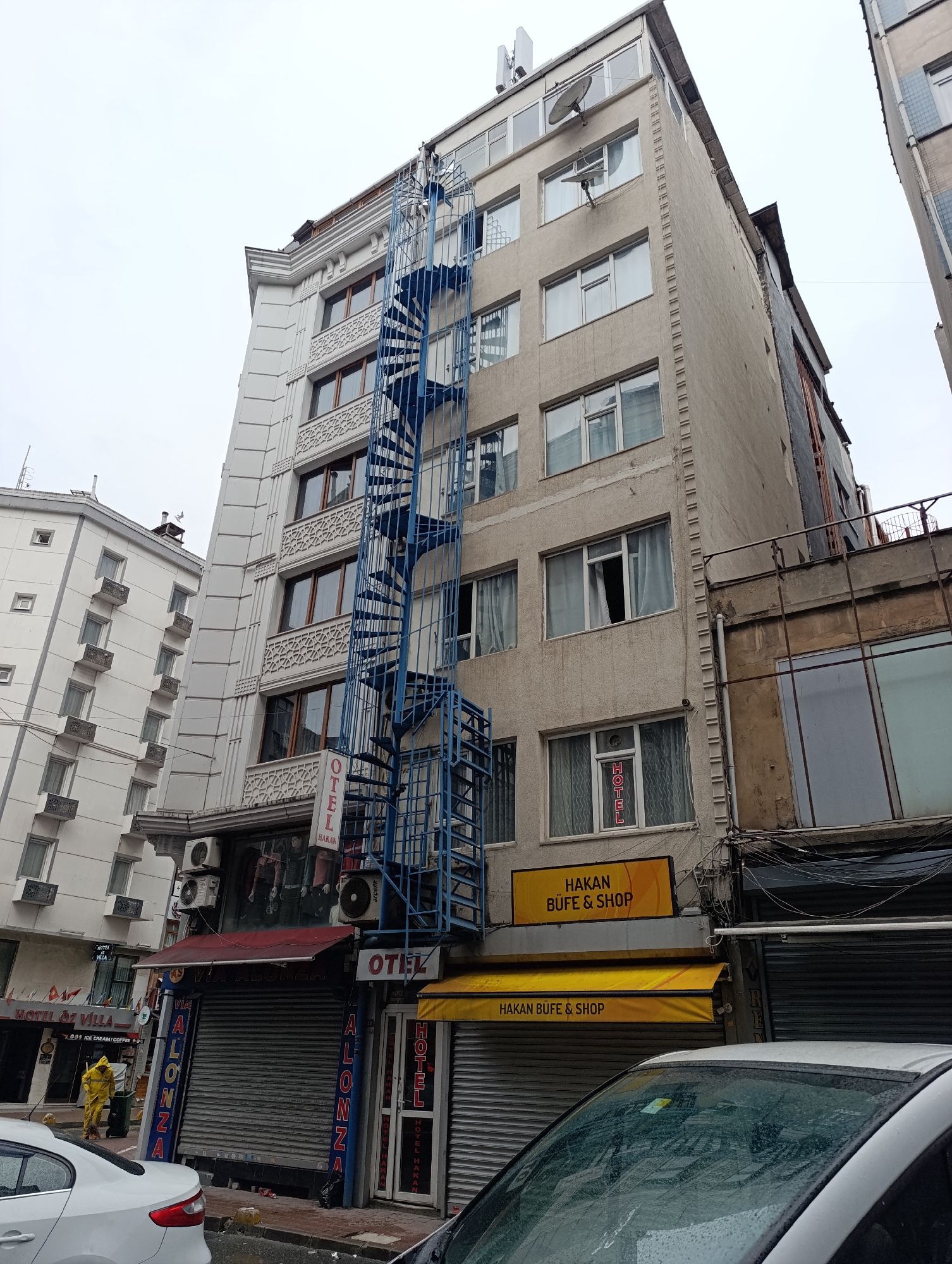 The image shows a street scene in Istanbul, Turkey, with a view of several buildings. One building has a blue spiral staircase on its exterior. The building's facade is unkempt and weathered. Commercial establishments on the ground level have closed metal shutters. Signage above the storefronts identifies them as "HAKAN BUFE & SHOP" and "OTEL." A partially visible car is parked in the foreground, with raindrops on its windshield. A person in a yellow protective suit is sweeping the sidewalk near another establishment named "HOTEL ÖZ VİLLA."
