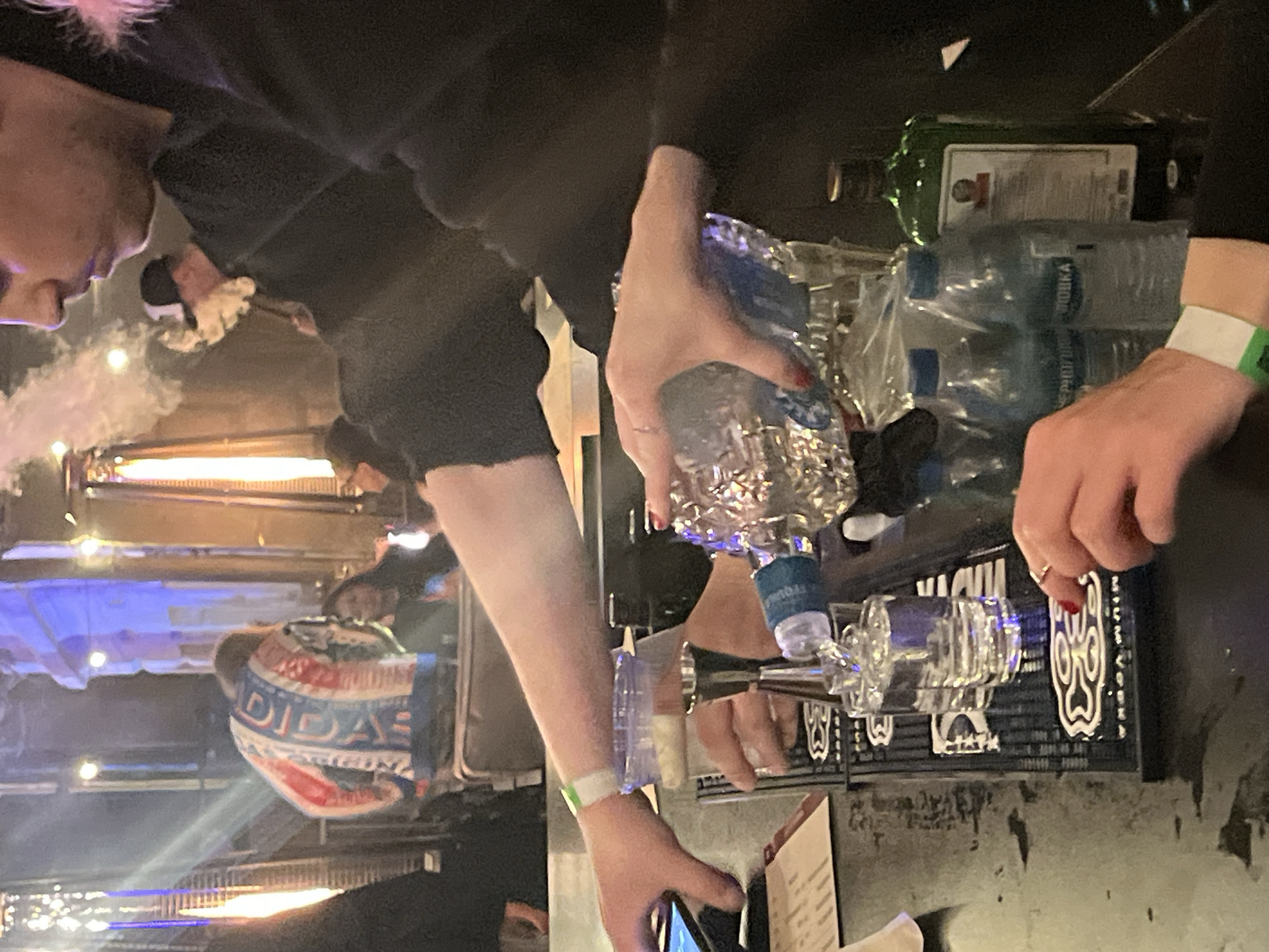 The image shows a close-up of a bar scene, focusing on the hands of a bartender and a customer. In the foreground, a bartender's hand is pouring a clear liquid from a decorative glass bottle into a shot glass. Another shot glass is already filled. The bartender is wearing a black long-sleeved shirt and a white wristband. A customer's hand, with red nail polish and a ring on their finger, is visible reaching towards the shot glasses. In the background, a person wearing a colorful jacket is visible, and there are lights and a hazy atmosphere, suggesting a nightclub or bar environment. Bottles of water and a green bottle of what appears to be alcohol are on the bar. The overall scene is one of drink preparation and service.