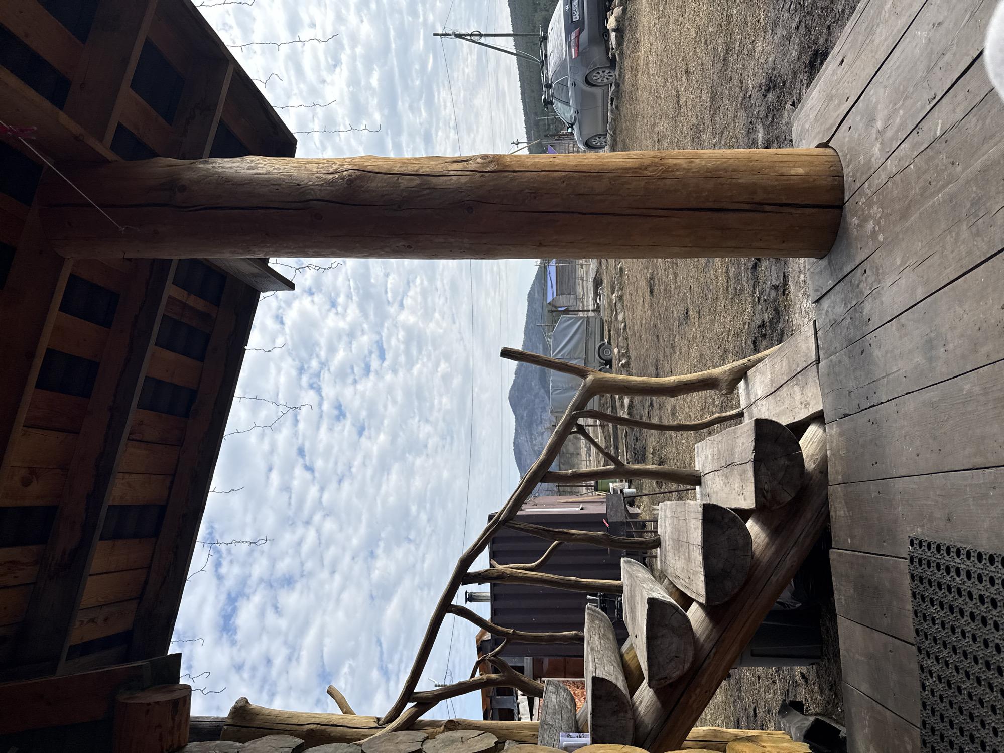 The image depicts a wooden structure, likely a cabin or outbuilding, in Tyulyuk, Russia. The structure features a log column supporting a roof and a wooden staircase with a rustic railing made from tree branches. Outside, a car and other structures are visible against a backdrop of a cloudy sky and a distant mountain. The ground is covered with sparse vegetation.