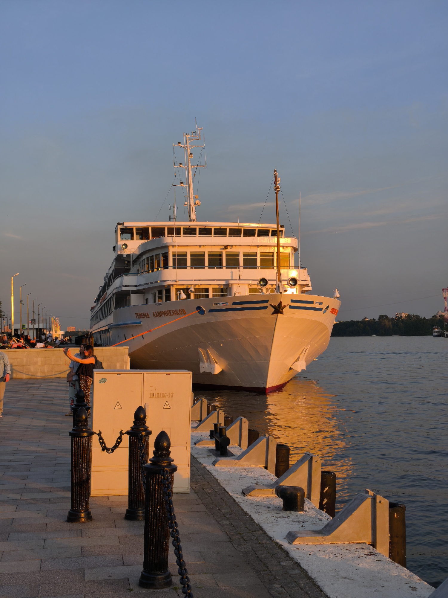 The image depicts a river embankment scene in Moscow, Russia, during the golden hour of late afternoon or early evening. A large, white passenger river cruise ship, identified by the red lettering "ГЕНЕРАЛ ЛАВРИНЕНКОВ" (General Lavrinenkov) on its side, is prominently docked at the concrete embankment, angled towards the viewer. The ship features multiple decks, numerous rectangular windows, and a white mast structure with various antennas at its apex. A red star emblem is visible on the bow.
The sun's low angle casts a warm, golden light across the scene. The ship's bow, the water, and the upper surfaces of the embankment infrastructure are strongly illuminated with an orange hue. The river water reflects this golden light.
On the paved embankment in the mid-ground, two women are engaged in taking a selfie with a smartphone, positioned near a light-colored utility box. This box bears the inscription "НИИК-17" and two black triangular warning symbols. Black, fluted bollards with decorative tops and chains segment the promenade area. Further down the embankment, to the left, several other people are visible, some seated or standing, appearing as silhouettes against the receding light. The far left also shows parts of a streetlight pole.
Along the water's edge, concrete mooring structures with black bollards are visible. The sky above is a gradient, transitioning from light blue at the top to a faint orange and pink near the horizon. In the distant background, across the river, treelines, a few buildings, and a tall red and white structure (possibly a tower or crane) are faintly discernible.