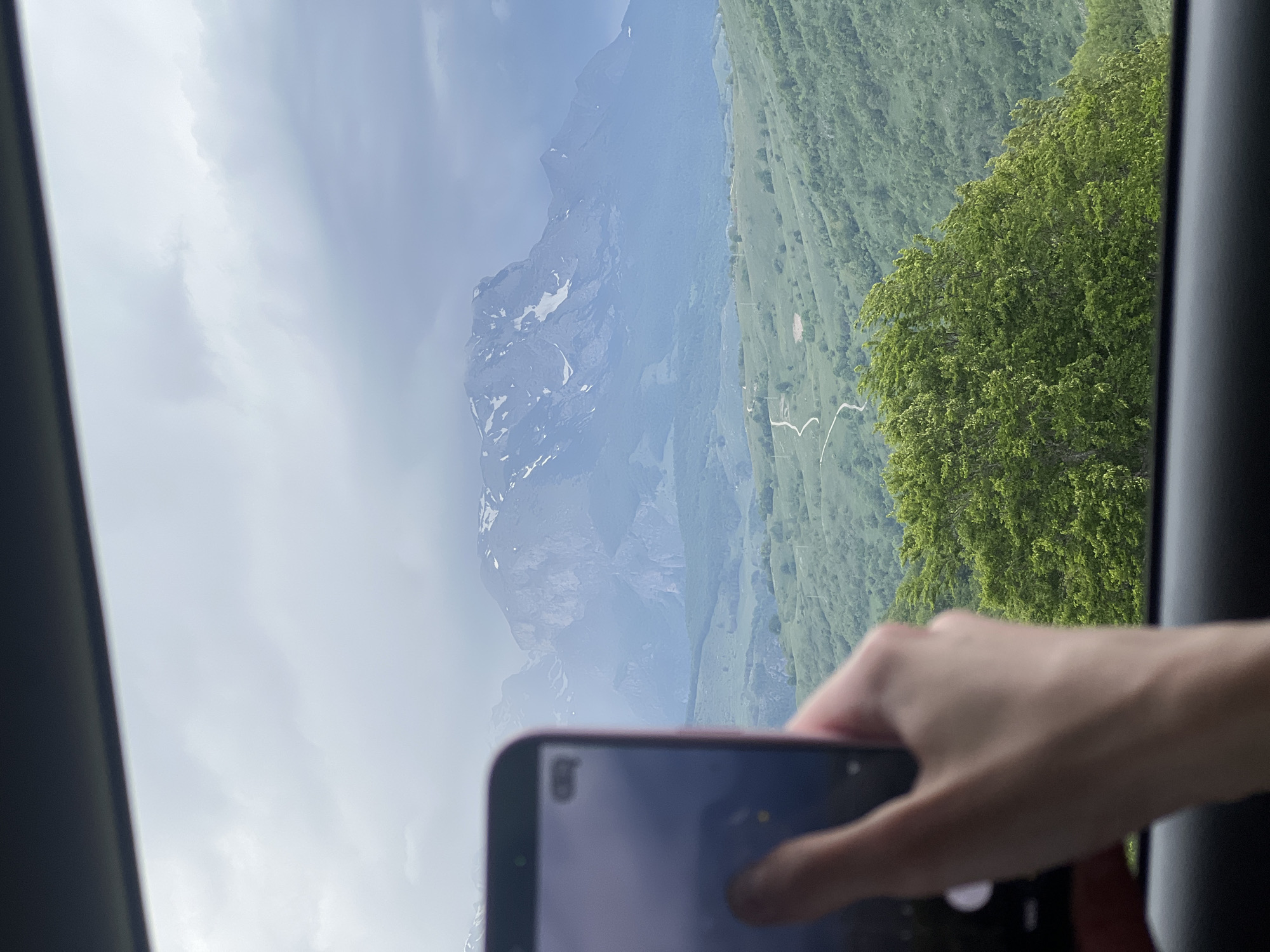 The image depicts a mountain landscape as viewed through a vehicle window. The foreground includes a hand holding a smartphone, partially obscuring the view. Through the window, a mountain range is visible with areas of snow cover. Adjacent to the mountains is a green, sloping hillside dotted with trees. It appears to be in the city of Pišče, Montenegro. The sky is overcast, indicating cloudy weather conditions. The presence of the window frame suggests the photograph was taken from inside a car or bus.
