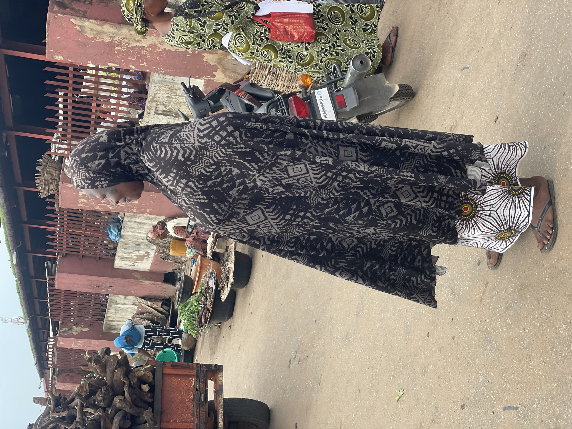 This image captures a scene at a market in Lokoja, Nigeria. In the foreground, a woman wearing a black and grey patterned headscarf and a black and grey patterned wrapper is standing and looking to her left. She is also wearing a white and patterned skirt underneath. To her right, another woman in a green and black patterned dress is visible, carrying a red bag. Behind them, a motorcycle is parked. The background reveals market stalls with various goods, including what appears to be firewood on a truck to the left and baskets of produce. The environment suggests a bustling, outdoor market with a dirt or paved ground.
