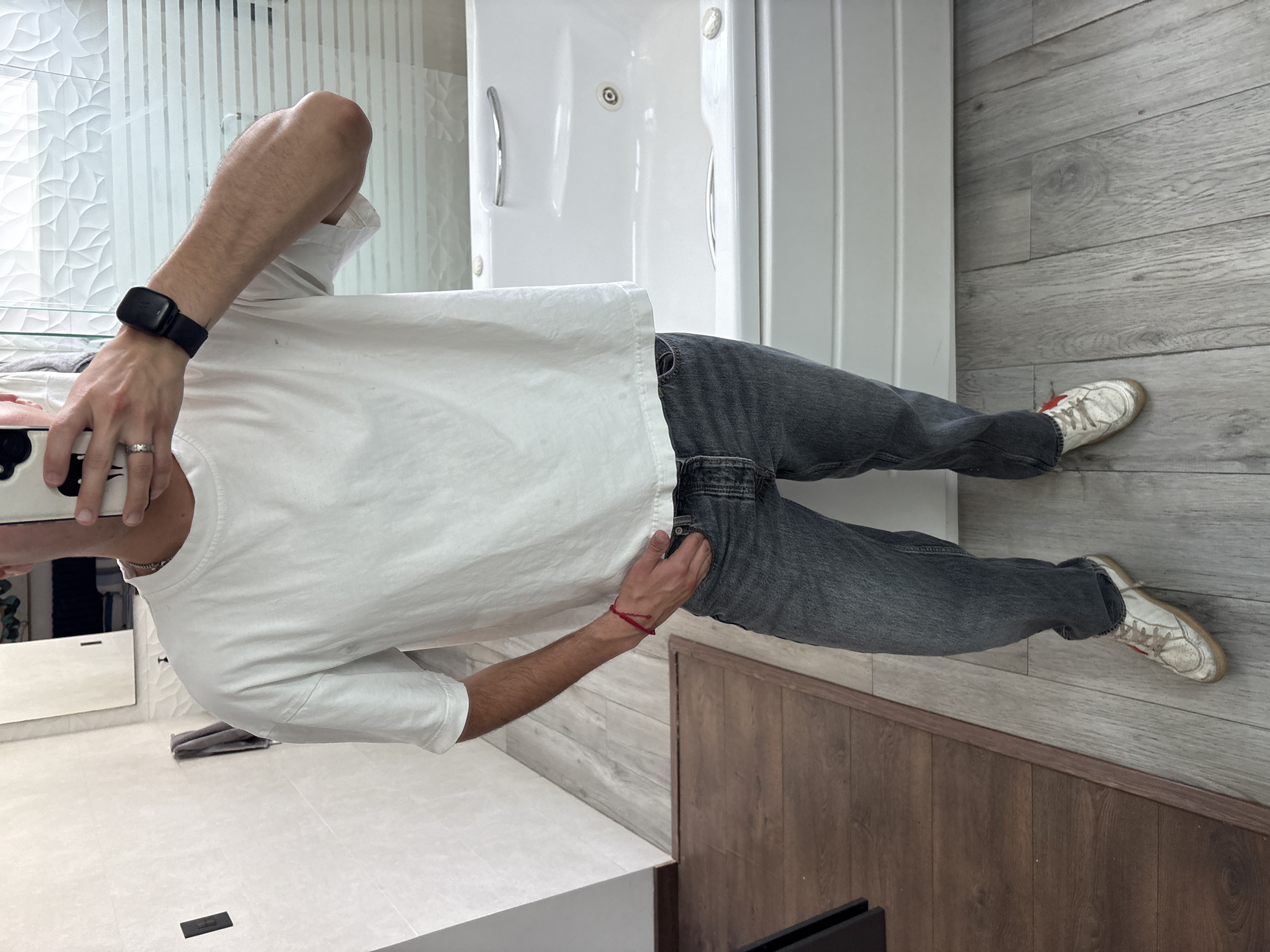 A portrait-oriented image, originally rotated 90 degrees clockwise, shows a person (likely male, based on visible arm hair) standing in a modern bathroom, possibly taking a mirror selfie. The person is cropped above the chest, with their head not visible. They wear a plain white short-sleeved t-shirt and dark gray, possibly washed-out black, jeans. White sneakers with red detailing and worn soles are on their feet.
On the left wrist, a black smartwatch or fitness tracker is visible, and on the right wrist, a red string bracelet is worn. A silver ring is on the left ring finger. The person holds a white smartphone with a black patterned case in their left hand, while their right hand is casually tucked into the front pocket of their jeans.
The bathroom features a mix of flooring and wall coverings. The immediate foreground has white tiled flooring, with a dark rectangular object (possibly a floor drain cover) near the bottom edge. To the right, the floor transitions to light gray wooden-look planks, which also extend up the wall. A dark brown wooden-look paneling runs along the base of the wall on the right. In the background, a white shower or bathtub enclosure with chrome handles and fixtures is visible. A reflection in a mirror captures part of the person's white t-shirt and the white vanity area. The lighting is bright and even, suggesting an indoor setting during the daytime. No discernible text is visible.
