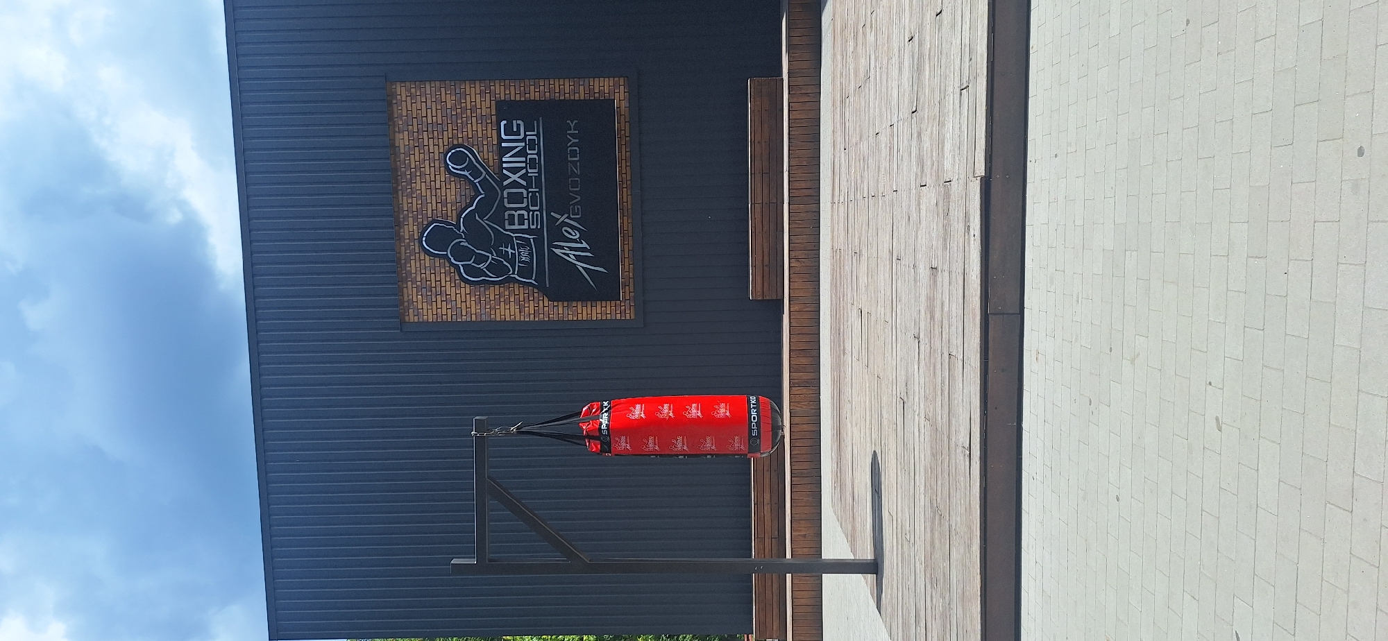 The image shows the exterior of a boxing school on a sunny day. The building features a dark, corrugated metal facade with a sign that reads "ALEX BOXING SCHOOL GVOZDYK" above a graphic of a boxing glove. A red punching bag hangs from a metal stand in front of the building, which appears to be a commercial establishment. The scene is set against a bright blue sky with some clouds, suggesting fair weather. To the right of the building, there is a paved area made of light-colored rectangular stones, and a section of wooden decking. The location is identified as Bila Tserkva, Ukraine. There is no activity or people visible in the image.