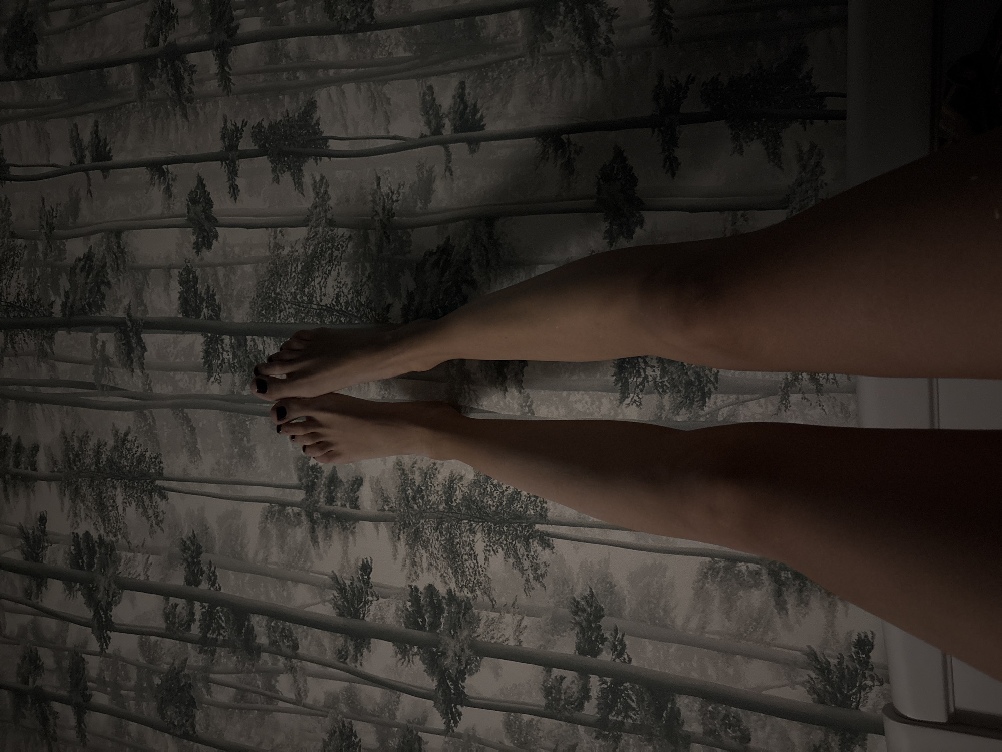 The image displays a close-up, top-down view of a person's legs and feet, posed against a backdrop of wallpaper patterned with trees. The lighting is dim, casting shadows that obscure some details of the legs. The person's toenails are painted black. The wallpaper features a stylized forest scene with tall, slender trees and dense foliage rendered in shades of grey and white, creating a somewhat abstract, textured effect. The lower portion of the image shows the edge of a white headboard. The scene is devoid of any specific indicators that would allow for its location to be identified as Krasnoyarsk, Russia.