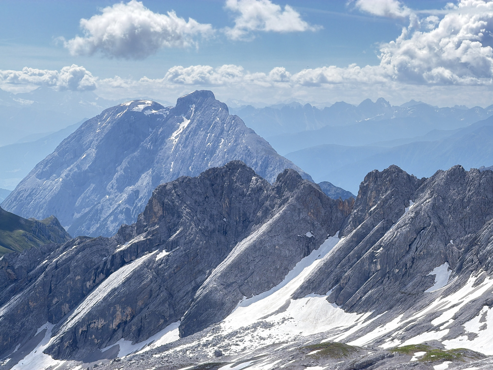 This is a landscape photograph taken from a high vantage point, likely a mountain peak, overlooking a vast mountain range. The foreground is dominated by the rugged, grey slopes of a mountain, with patches of snow clinging to its steep inclines. These slopes are characterized by sharp, jagged rock formations and a sense of dramatic elevation.
In the middle ground, a prominent, snow-capped mountain peak rises majestically, its summit distinct against the clear blue sky. This peak appears to be the highest point in the immediate vicinity.
The background reveals a seemingly endless expanse of progressively bluer, hazy mountain ranges, fading into the distance. These distant peaks suggest a much larger mountain chain. The sky above is a vibrant blue, dotted with fluffy white cumulus clouds, some of which are large and billowy, casting soft shadows.
The scene is one of immense natural beauty and scale, emphasizing the grandeur of the alpine environment. The lighting suggests it is daytime, with sunlight illuminating the mountain faces and highlighting the textures of the rock and snow. There are no people or man-made structures visible in the image, focusing solely on the natural landscape.
This scene is likely representative of the mountainous region surrounding Garmisch-Partenkirchen, Germany, known for its dramatic alpine scenery and peaks like the Zugspitze, Germany's highest mountain, which could be depicted in the background.