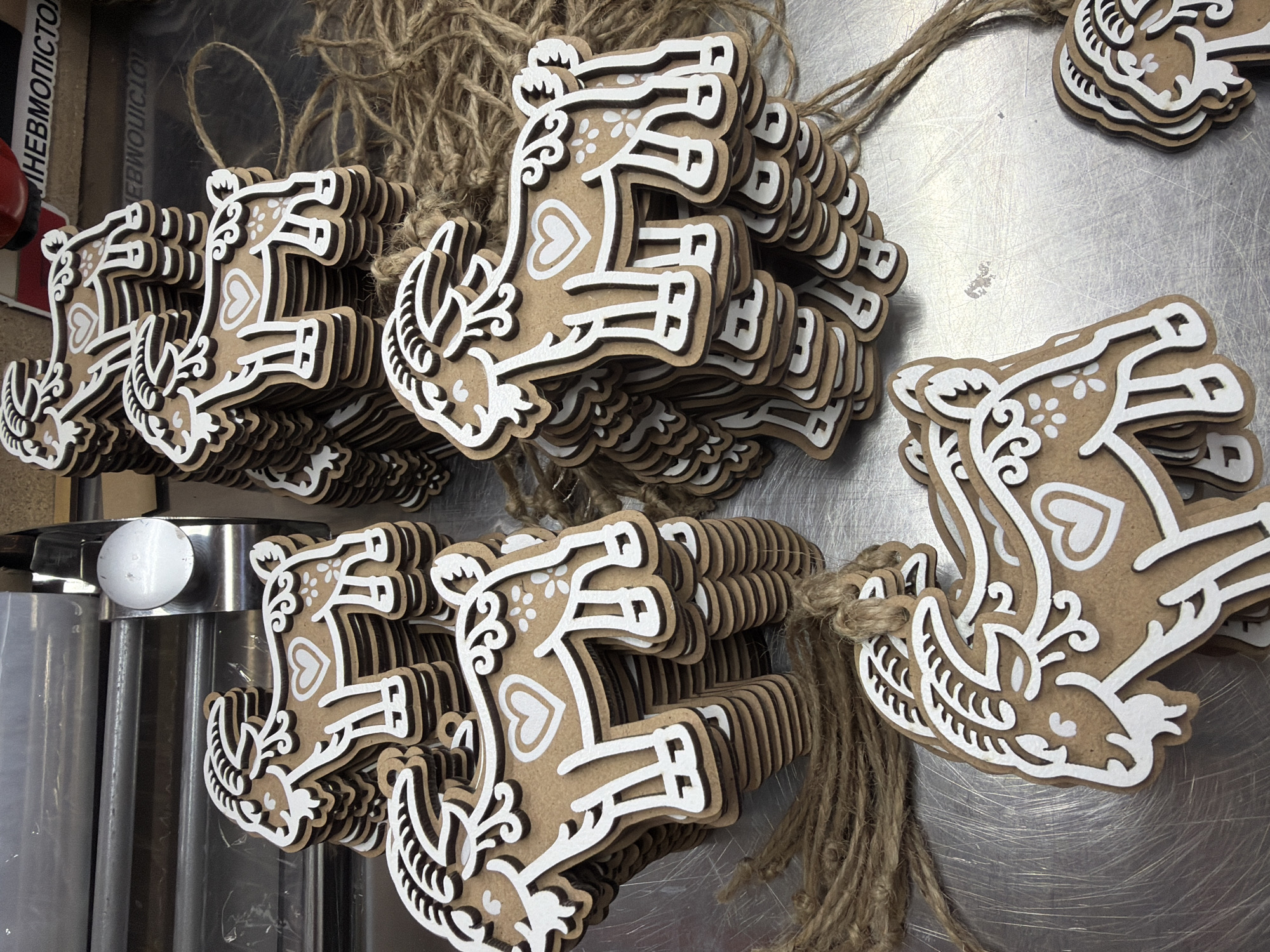 The image shows a collection of stacked wooden goat-shaped ornaments. The ornaments are made of light brown wood with white decorative cutouts depicting a heart on the side, floral patterns, and facial features. Each ornament is layered, creating a multi-dimensional effect. Some of the ornaments are tied together with rough twine. The ornaments are arranged in stacks on a metallic, possibly stainless steel, surface. In the background, there is machinery that appears to be part of a manufacturing or production line, with rollers and metallic components visible. There is also some text on a cardboard backing in the far background, which is not entirely legible. The lighting suggests an indoor setting, likely a workshop or production facility.
