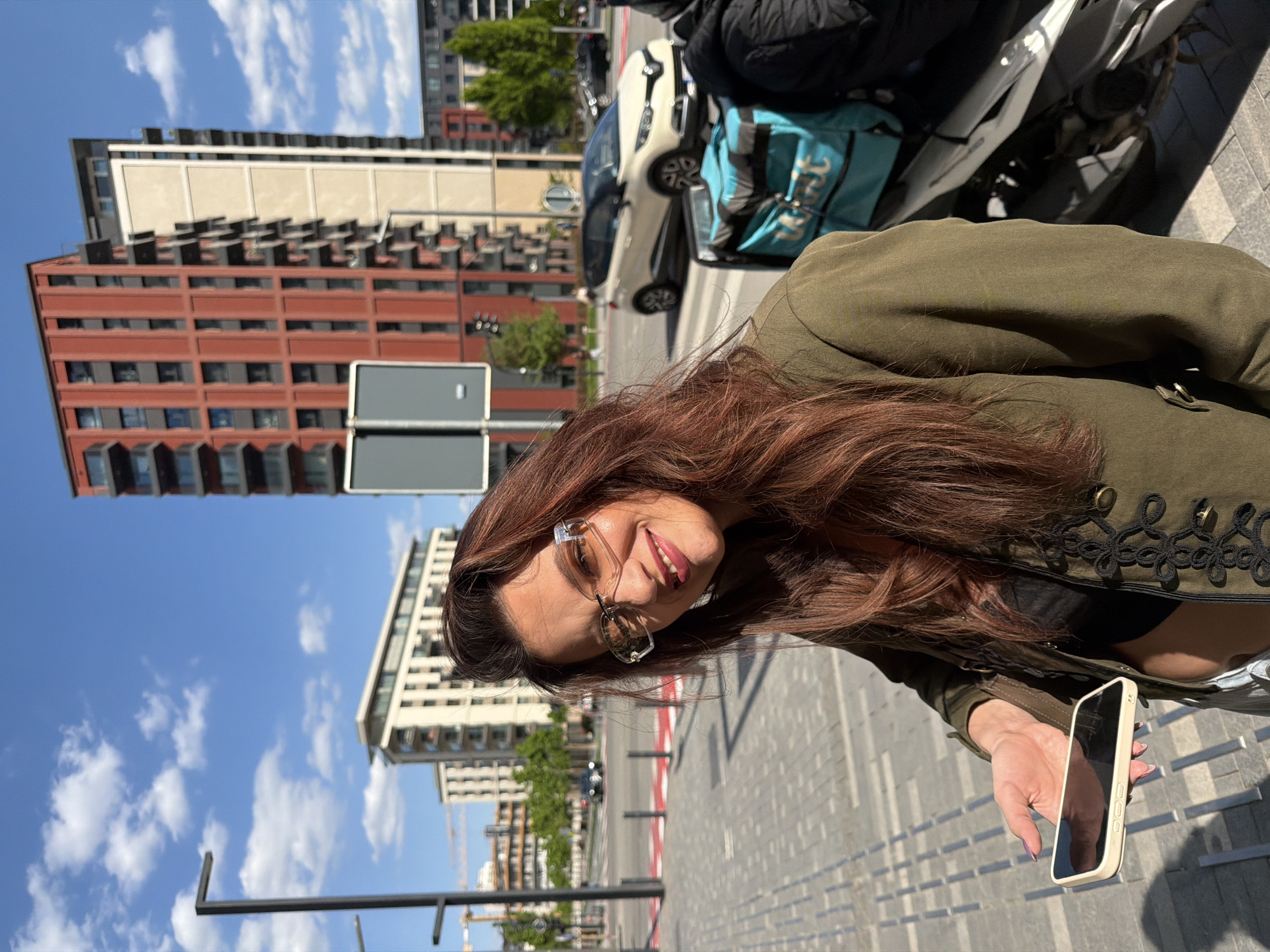 An outdoor portrait captures a smiling woman against an urban backdrop in Belgrade, Serbia. The woman, with long, wavy brown hair, is positioned on the right side of the frame, looking towards the viewer. She wears sunglasses, a dark green jacket with black decorative embroidery on the lapels, and a black top. In her right hand, she holds a light-colored smartphone.
The setting is a city street or paved area, bathed in bright daylight under a clear blue sky with scattered white clouds, indicating sunny weather, possibly mid-day or afternoon due to strong shadows. In the background, several modern buildings are visible, including a prominent red-brown one with numerous rectangular windows and a lighter-colored, multi-story building. To the woman's right, behind her, a white car and a motorbike are parked, along with a bright blue delivery bag, possibly belonging to a food delivery service, with partially visible text that appears to be "WOLT". The woman's joyful expression, with a wide smile, suggests a pleasant and relaxed mood.