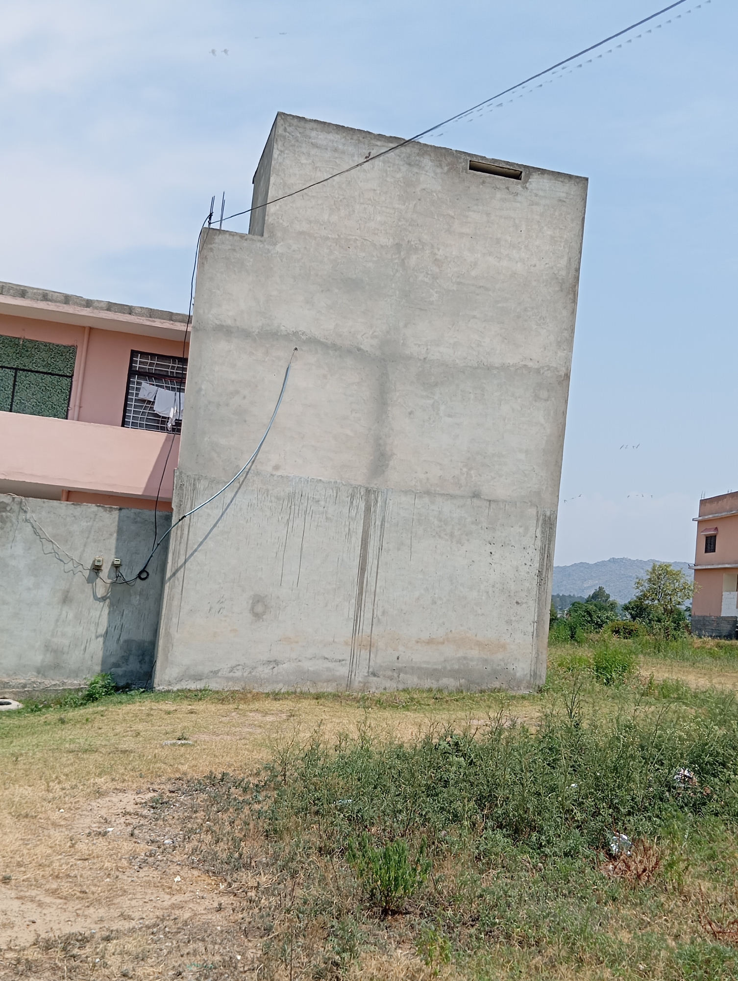 A tall, unfinished concrete structure leans dramatically to the right in the foreground. To the left of this structure is a lower, pink building with a green patterned window and a window with a metal grate. Several white articles of clothing are hanging on a clothesline behind the metal grate. A dark wire runs from the top of the concrete structure down to electrical meters attached to a grey wall. The ground in front of the structures is dry grass and dirt, with some weeds and small stones. In the background, a tan building with a brown roof is visible, as are green trees and distant, hazy mountains under a light blue sky with some small, distant birds.