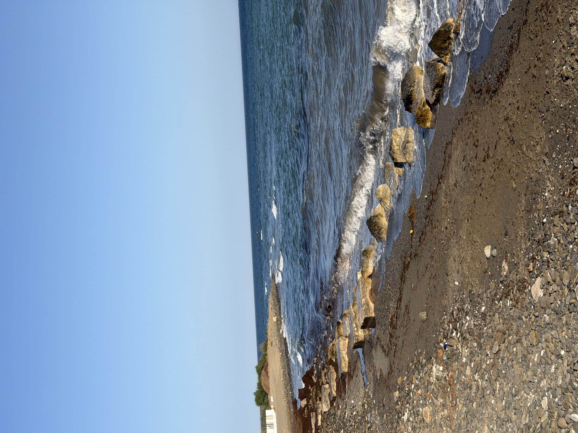 The image depicts a rocky beach with the Mediterranean Sea in Saladillo-Benamara, Spain. The sky is clear and blue. The foreground is a mix of dark pebbles and sand, with several large, square, light-colored concrete blocks partially submerged in the shallow water or resting on the shore. Beyond these, the sea is a deep blue, with moderate waves crashing against the shore, creating white foam. Some smaller, rounded rocks are scattered along the water's edge. In the distance, a small pile of dirt and some green vegetation are visible on the shore, along with a white fence-like structure. The overall scene is that of a natural coastal environment with some man-made elements.