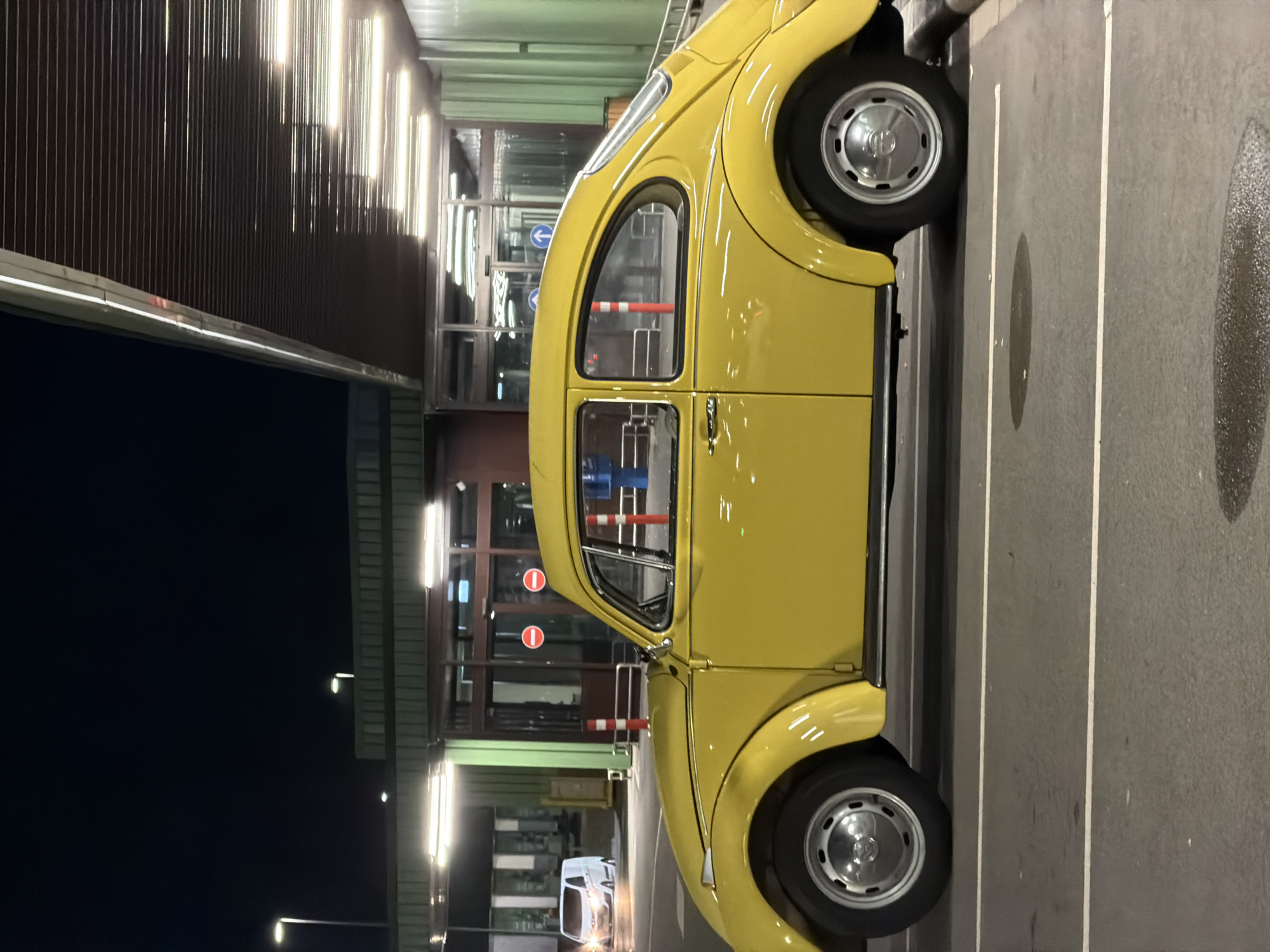 A yellow Volkswagen Beetle is parked at night in what appears to be a covered parking area, likely at a public facility or transit hub in Leudelange, Luxembourg. The car is positioned in a parking spot marked by white lines on a dark asphalt surface. The building behind the car features a modern, angled roof with embedded lighting, and large glass doors or windows. The overall lighting suggests it is nighttime, with artificial lights illuminating the scene. In the background, a white van is visible, also in a parking area. The environment is clean and well-maintained, characteristic of a public urban space.