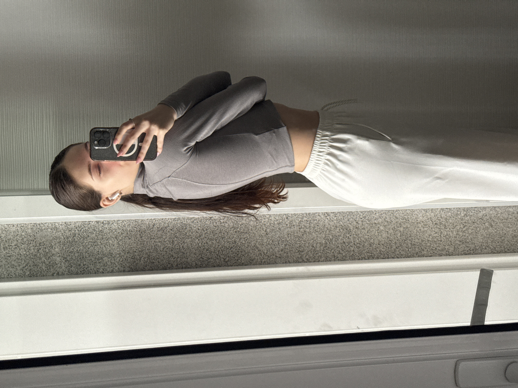 This mirror selfie, taken in Sofia, Bulgaria, features a young woman standing and capturing her reflection indoors. She has long, dark brown hair that extends past her shoulders. The woman is wearing a grey, long-sleeved crop top which reveals her midriff, and loose-fitting white pants with a visible drawstring at the waist. White wireless earbuds are visible in her ears.
She holds a black smartphone in her right hand, positioned to take the selfie. The phone case is black with a distinct white symbol: a circle with a diagonal line through it, resembling a "peace sign." Her face is partially obscured by the phone, but her left eye and nose are visible. Her fingernails are painted with a light pink polish.
The background, reflected in the mirror, shows a light grey wall with subtle vertical textured lines. Below the wall, there is a darker grey speckled surface, possibly a granite windowsill or ledge, followed by a white horizontal frame at the bottom of the image. Bright, natural light appears to be entering from the left, creating soft shadows and highlighting parts of the scene, suggesting it is daytime. The image conveys a casual and relaxed atmosphere.