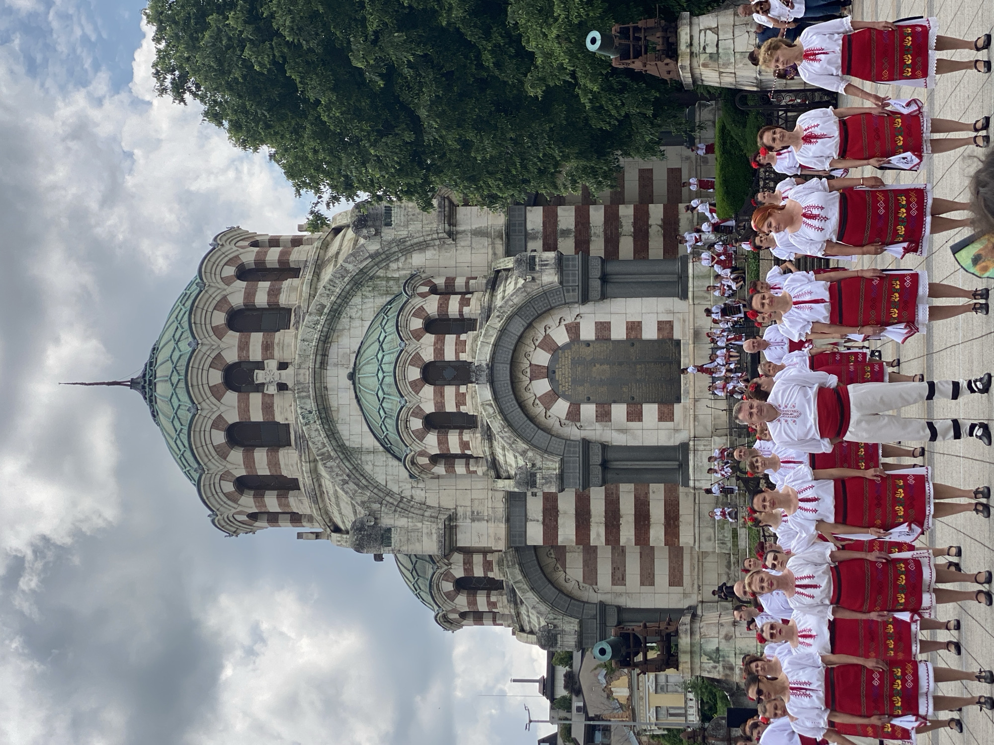 The image shows a group of people in traditional Bulgarian folk costumes performing in front of the St. George the Victorious Chapel Mausoleum in Pleven, Bulgaria. The mausoleum is a large, ornate building with a green dome, visible on the left side of the image. The performers are lined up in rows, wearing white shirts with red embroidery and red skirts with patterned borders. Some hold drums. A cannon is visible near the building. The sky is overcast.