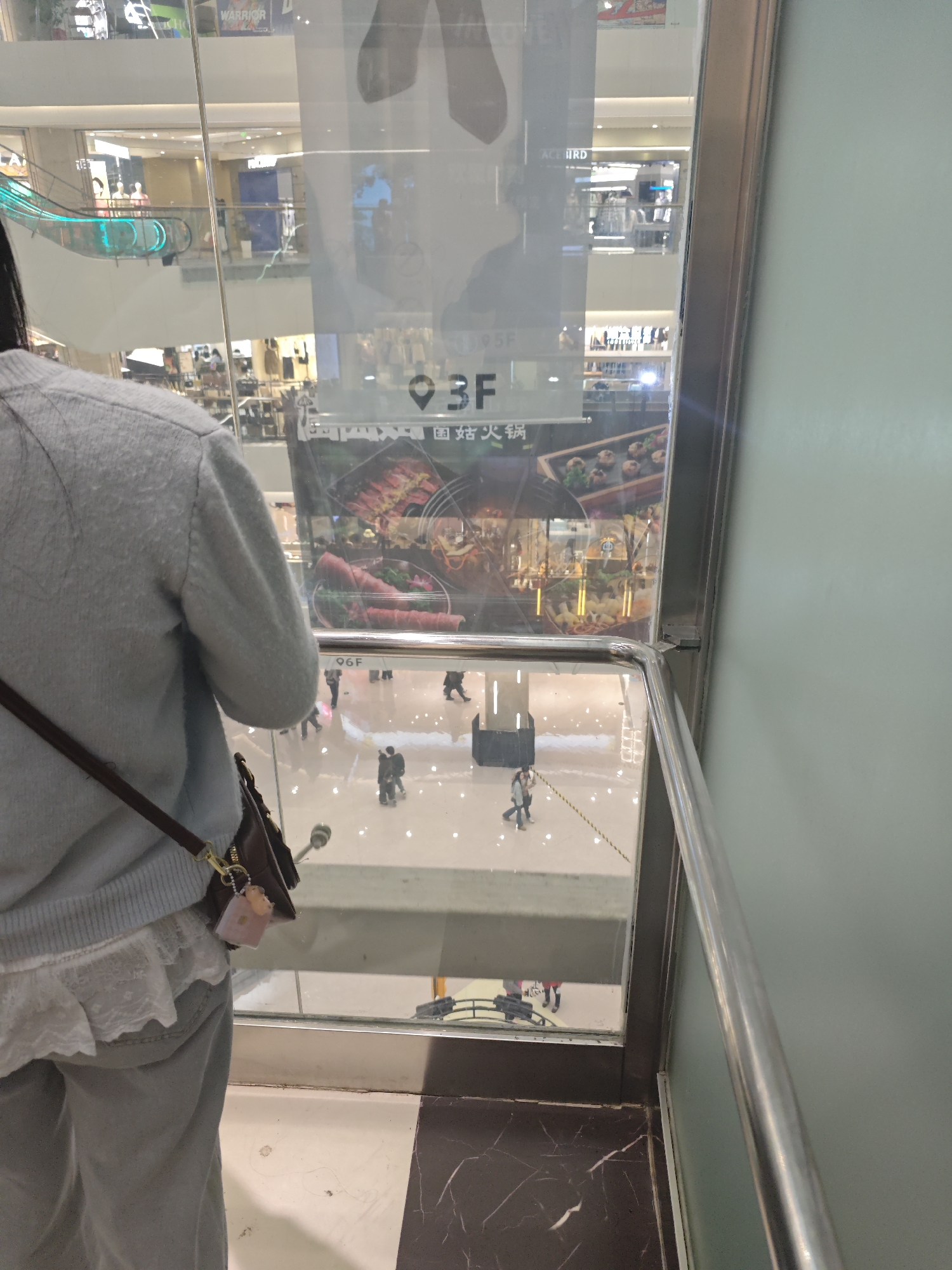A person is standing in an elevator looking out at a shopping mall in Liangjiang, China. The mall has multiple floors, visible through the elevator window, with advertisements for restaurants and shops. People can be seen walking around on the floors below. The elevator's interior includes a railing and marble flooring. The person in the elevator is wearing a light blue sweater, gray pants, and has a brown purse hanging on their shoulder.