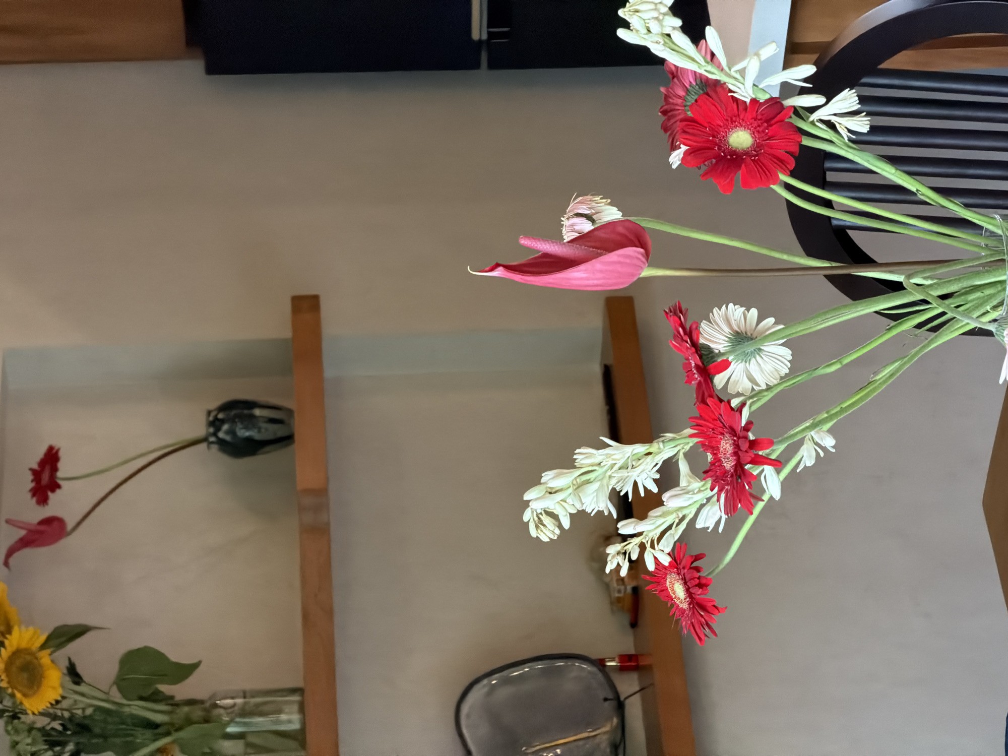 The image shows an indoor scene featuring a floral arrangement with red gerbera daisies and white tuberoses prominently displayed, set against a backdrop of beige walls and wooden shelves. Another floral arrangement with sunflowers is visible on a lower shelf. The scene appears to be in a residential setting with furniture visible in the background, including a black chair or stool. Without additional information, the specific location in Mengwi, Indonesia cannot be determined.