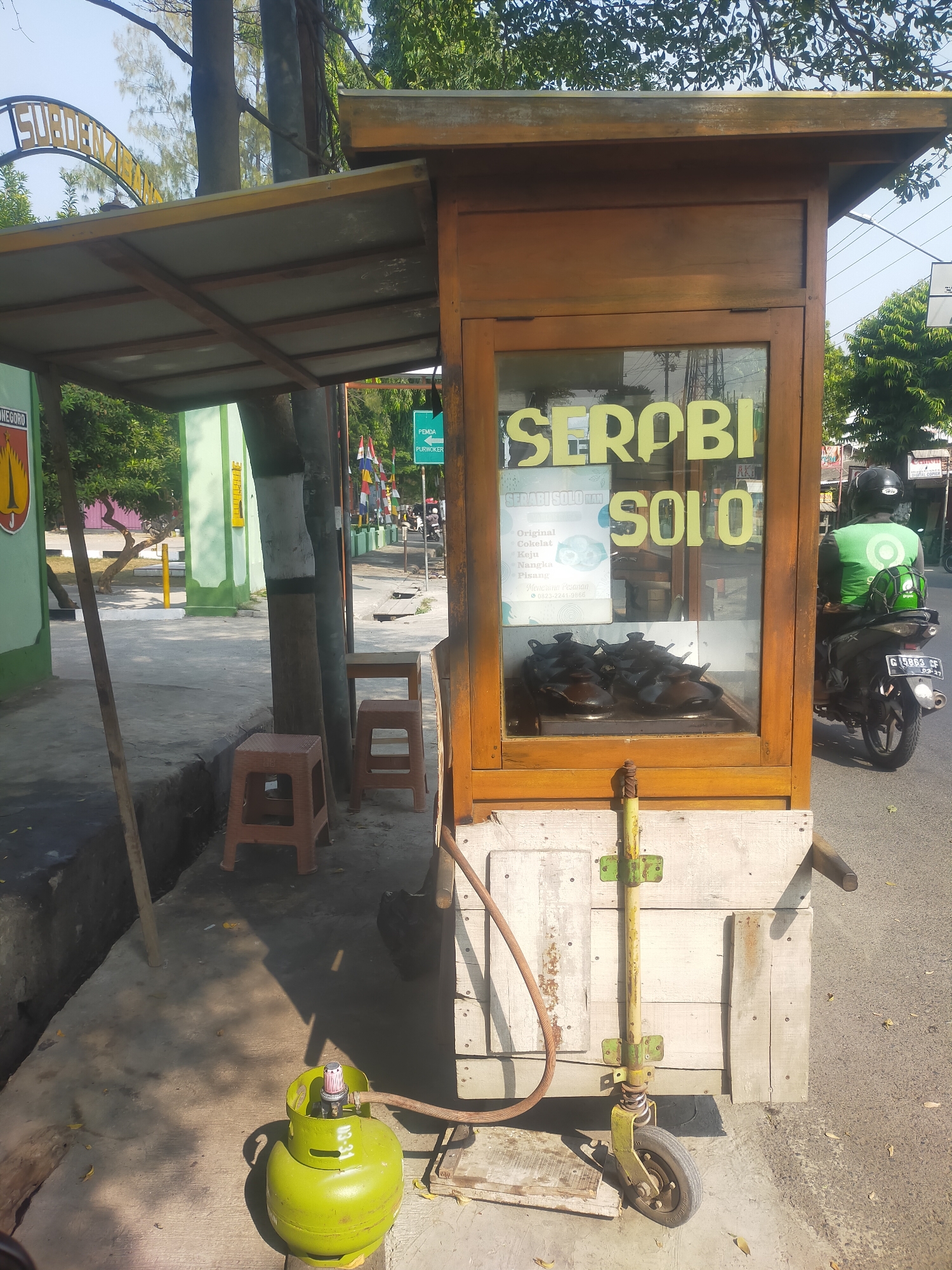 The image shows a street vendor stall selling "Serabi Solo" in Slawi, Indonesia. The stall is constructed of wood and has a glass display case where the serabi is being cooked in small, round pans. The words "SERABI SOLO" are visible in large yellow letters on the front of the stall. A green gas cylinder is connected to the stall by a hose, indicating a cooking fuel source. In the background, a person wearing a green jacket with a logo and a black helmet is riding a motorcycle. The street is paved, and there are trees and buildings visible in the background. A sign pointing to "PEMDA PURWOKERTO" suggests the location is within proximity to the Purwokerto regional government office. There are also two small brown stools placed near the stall. The overall scene depicts a typical street food vending operation in an urban environment.