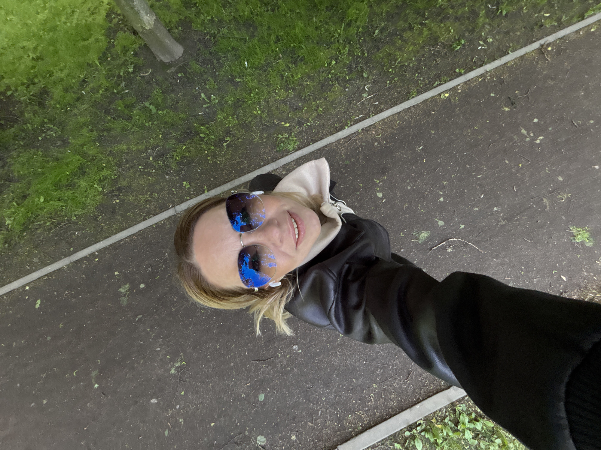 A woman takes a selfie. The woman has blond hair and wears reflective sunglasses, a black leather jacket, and a white sweatshirt. The reflection in her sunglasses appears to depict trees. She is on a paved walkway, and grass is visible on either side of the path. The environment is outdoors and appears to be in a park. Given that the location is specified as Saint Petersburg, Russia, it is reasonable to assume that this photo was taken in a park there.
