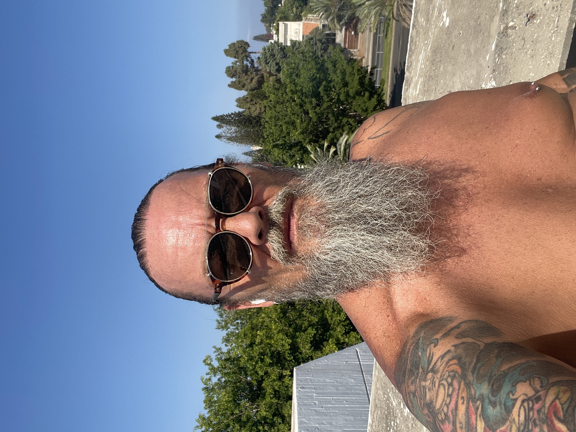 A shirtless man with a long grey beard and tattoos on his left arm and shoulder stands outdoors, looking at the camera. He is wearing sunglasses and white wireless earbuds. He has a piercing on his right nipple. Behind him is a clear blue sky, lush green trees, a building, and a road. The man is positioned on a concrete rooftop, which is visible in the foreground. The background suggests a urban or suburban setting in Tel Aviv, Israel.