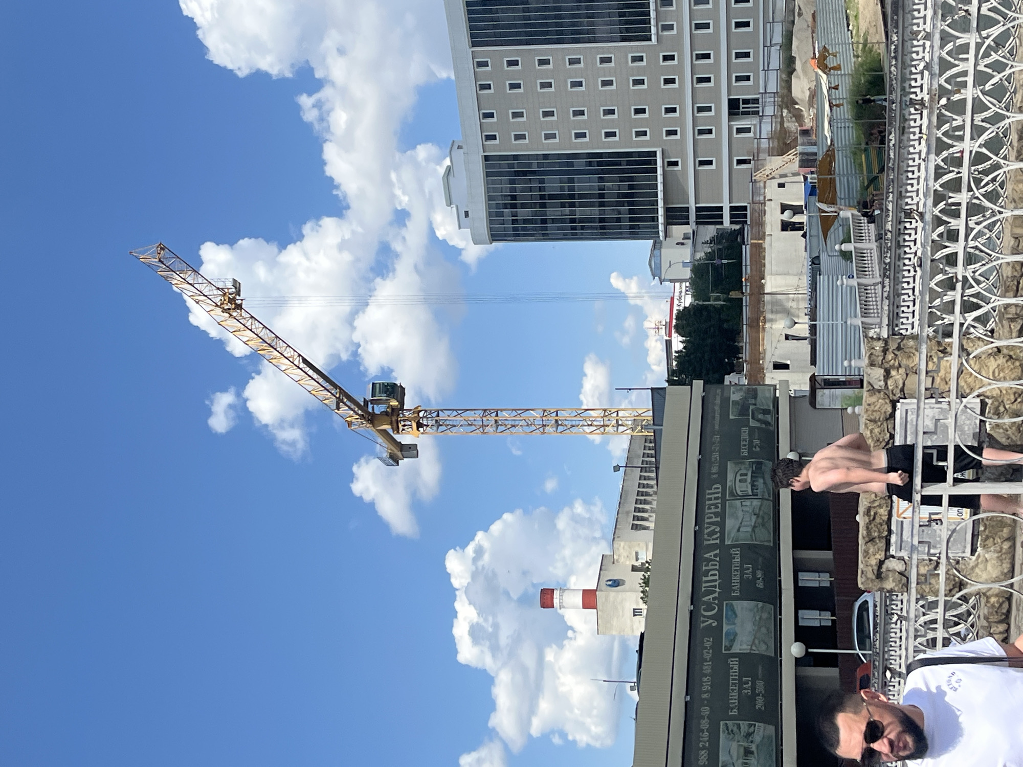 A yellow construction crane is prominent in the image, towering over a partially constructed building and a modern multi-story building with a glass facade. In the foreground, a man wearing sunglasses and a white t-shirt is visible, along with a younger shirtless man in black shorts. A decorative white metal fence runs along the bottom of the image, behind which is a stone embankment. The scene appears to be a waterfront area, possibly a park or public space, with a large billboard for "УСАДЬБА КУРЕНЬ" (Usadba Kuren), a banquet hall and event venue, visible on the left. The sky is blue with scattered white clouds. The image is likely taken in Krasnodar, Russia, given the signage.