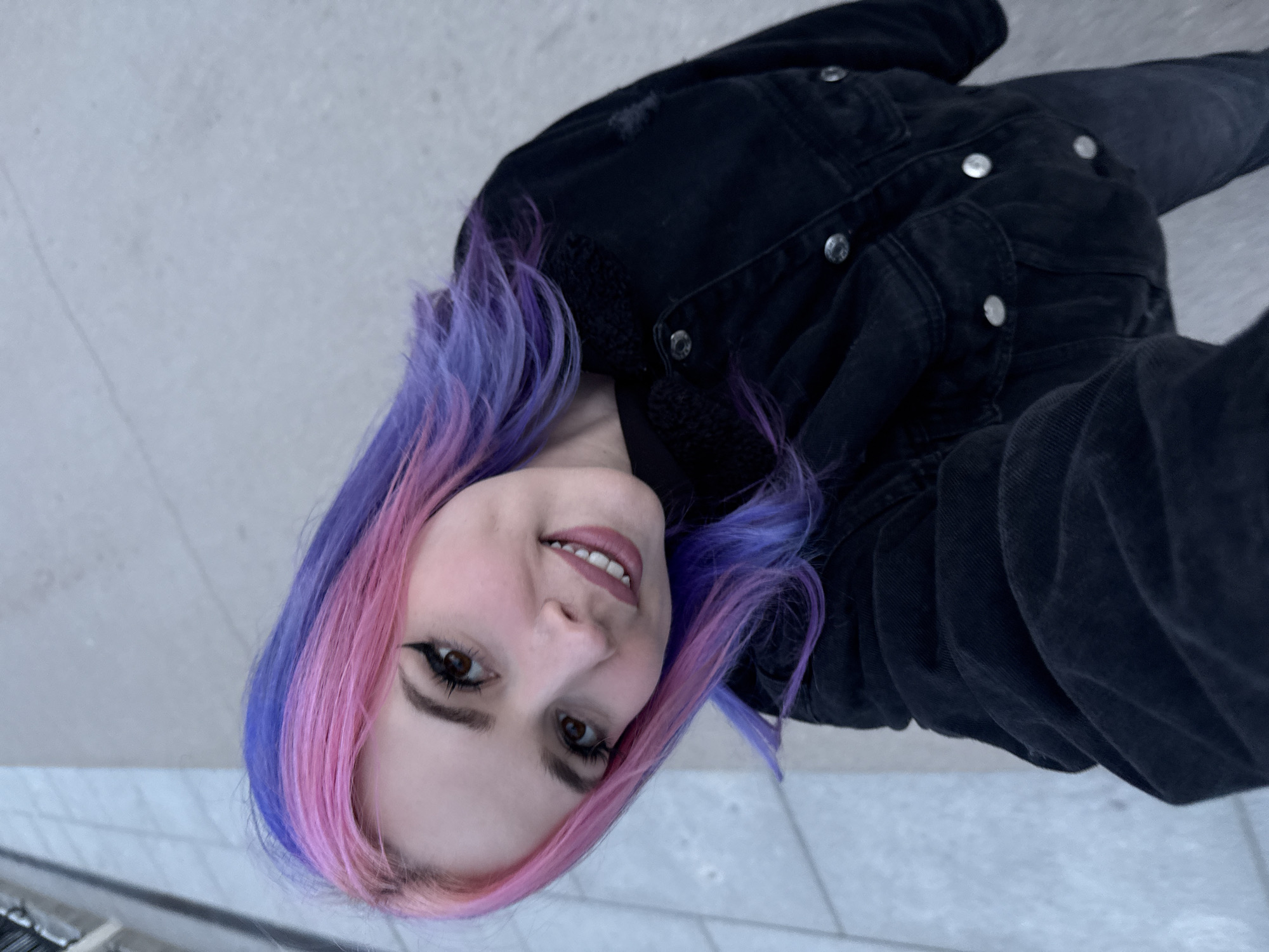 A young woman with dyed pink and purple hair is pictured in a selfie taken outdoors, possibly in the city of Madrid, Spain. She is smiling slightly at the camera and wearing dark eyeliner. She is wearing a dark-colored denim jacket with metal buttons and a sherpa collar. The background is a light gray color, which appears to be concrete or stone paving, potentially a sidewalk. The photo appears to have been taken from a low angle, looking upwards towards the subject.