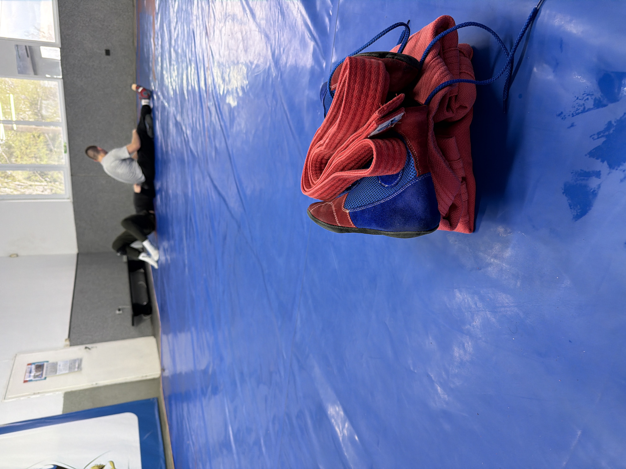 The image depicts an indoor sports facility, likely a wrestling or combat sports gym, located in Panagyurishte, Bulgaria. In the immediate foreground, a pair of wrestling shoes is prominently displayed on a textured blue mat. The shoes are distinctively red and blue, featuring blue laces, and a red fabric item, possibly a piece of clothing or a towel, is neatly folded beside them.
In the blurred background, two individuals are visible on the same blue mat, engaged in what appears to be a training session or practice. One person, wearing a light-colored t-shirt and dark pants, is in a bent-over posture, suggesting a grappling or wrestling stance, with the other person mostly obscured.
The room features white and grey walls, with large windows allowing natural light to illuminate the space, indicating it is daytime. On a grey wall section, a framed item is visible, likely a certificate or a poster, though any text on it is illegible due to the distance and blurriness. The blue mat, characteristic of wrestling areas, covers the floor where the activity is taking place. The overall scene captures a moment during a sports training session.