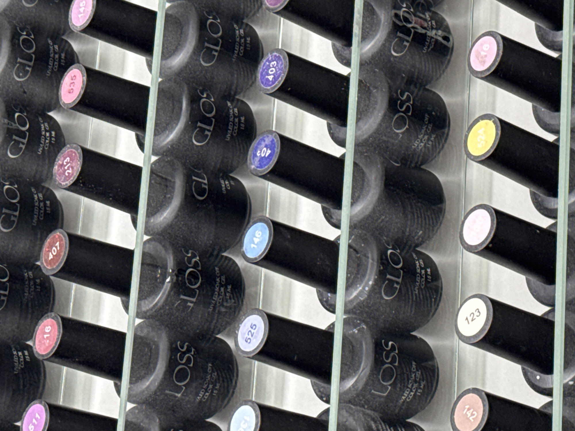 The image displays a tiered shelf holding numerous bottles of "GLOSS" brand UV-cured nail polish. The bottles are black with "GLOSS" and "UV/LED SOAK OFF COLOR GEL 15 ML" printed in white on their sides. The caps of the bottles have circular stickers with numbers indicating the shade. In the top row, visible numbers include 511 (pink), 516 (red), 407 (orange-red), 213 (pink), 535 (pink), and 145 (pink). The second row shows numbers like 351 (light blue), 525 (light blue), 146 (light blue), 403 (purple), and 403 (purple). The third row has numbers such as 142 (beige), 123 (white), and 524 (yellow), and 816 (pink). The image is a close-up shot, suggesting a retail or display setting, possibly a nail salon or beauty store. The lighting is bright, highlighting the products. The location is not specified in the image itself, however, assuming it is in Budva, Montenegro, this could be a retail display within a beauty supply store or a nail salon in the city.