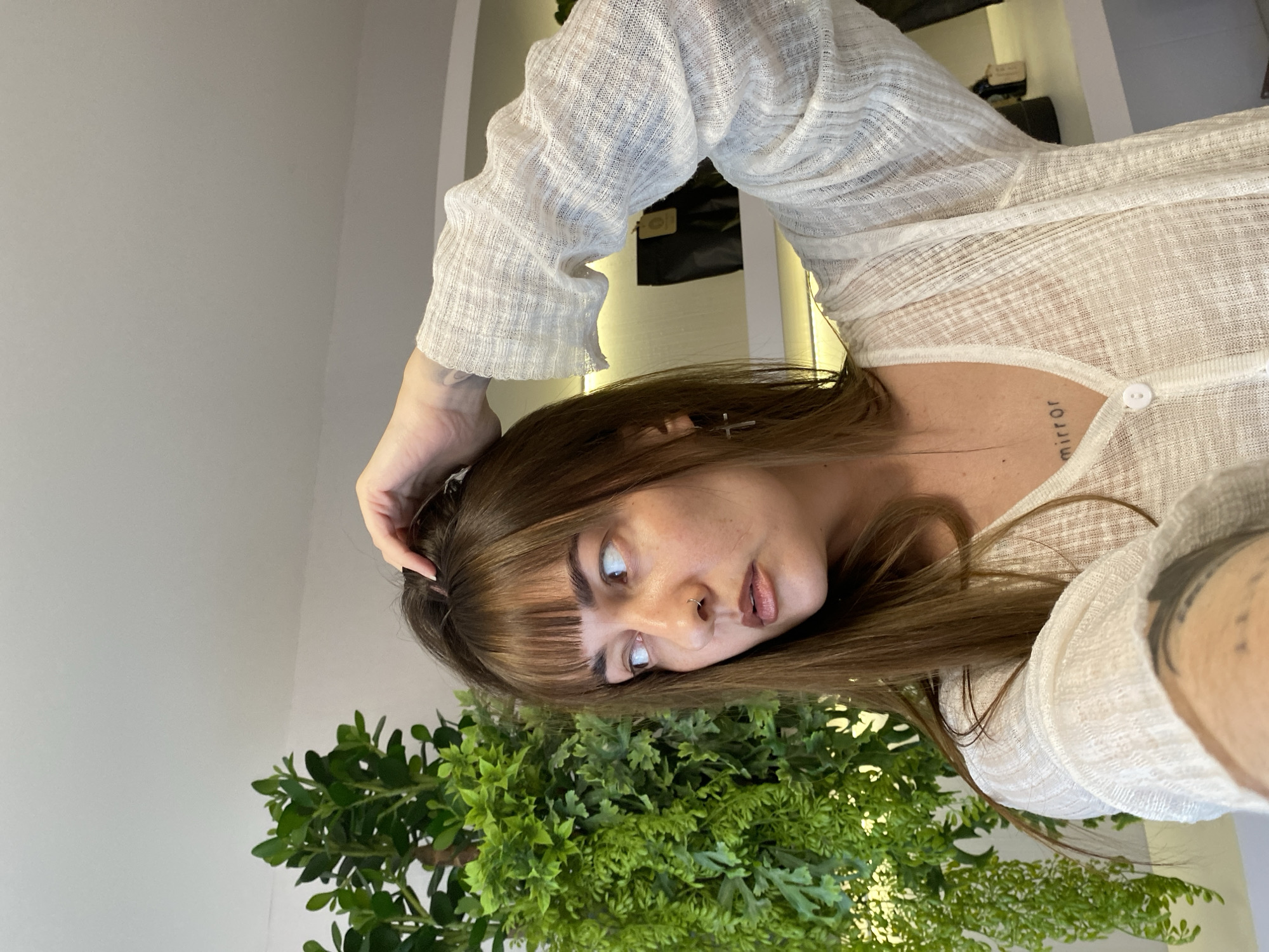 A woman with brown hair and bangs is looking upwards, with her right hand touching her hair. She is wearing a sheer, white, long-sleeved button-down shirt. Her right forearm is visible, showing a tattoo. A plant with green leaves is positioned near her face. She has a cross hair clip in her hair, a tattoo of the word "mirror" on her upper chest, and a nose ring. The background includes a white wall. There is no information to indicate that this image was taken in Votkinsk, Russia.