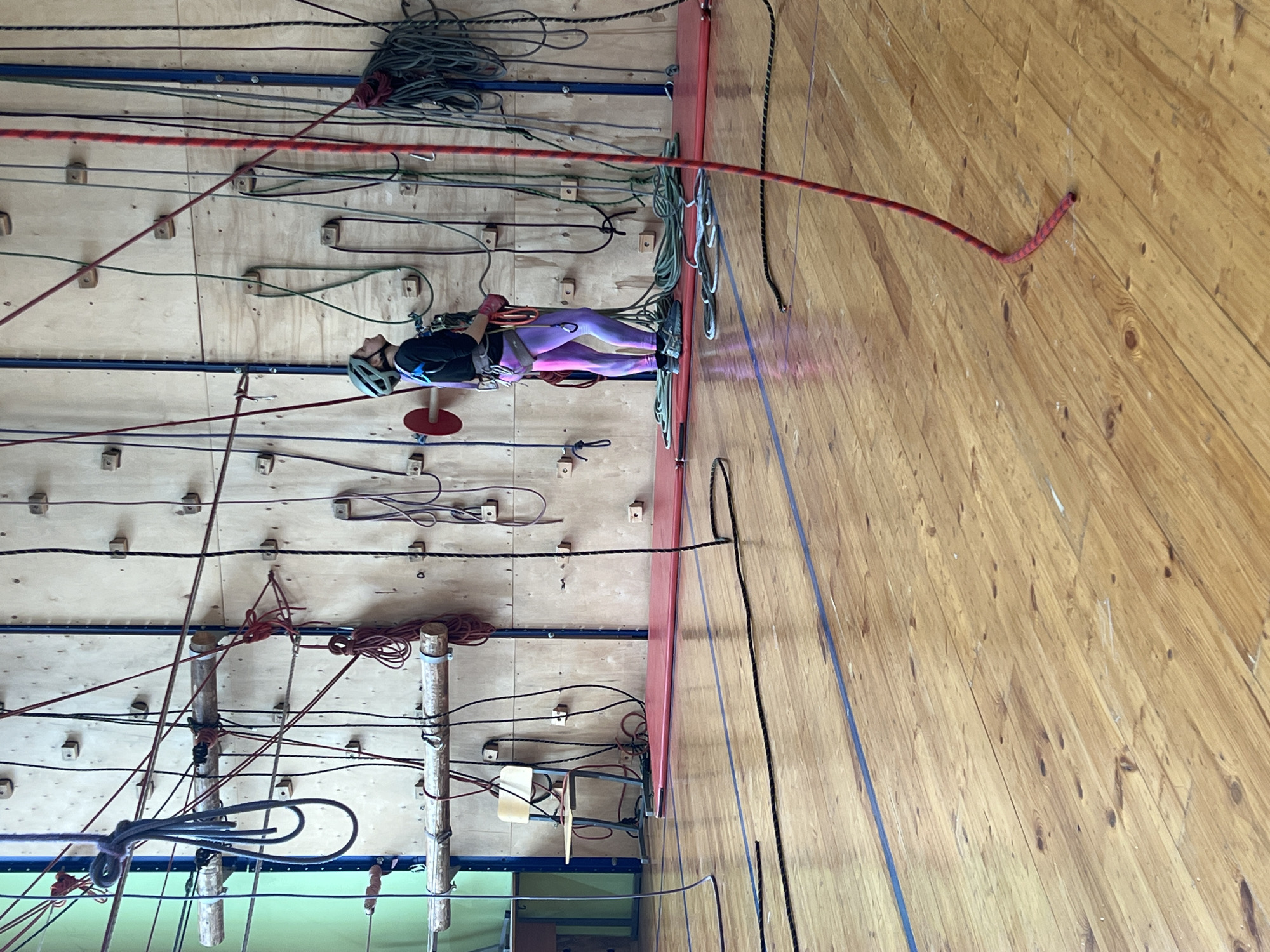 A person wearing a helmet, black shirt, and purple pants is suspended on a climbing wall. They are secured by ropes and harnesses. The wall is made of wood and has various handholds and ropes attached to it. The location is likely an indoor climbing gym, potentially located in the city of Chaikovsky, Russia. The environment is cluttered with climbing equipment.