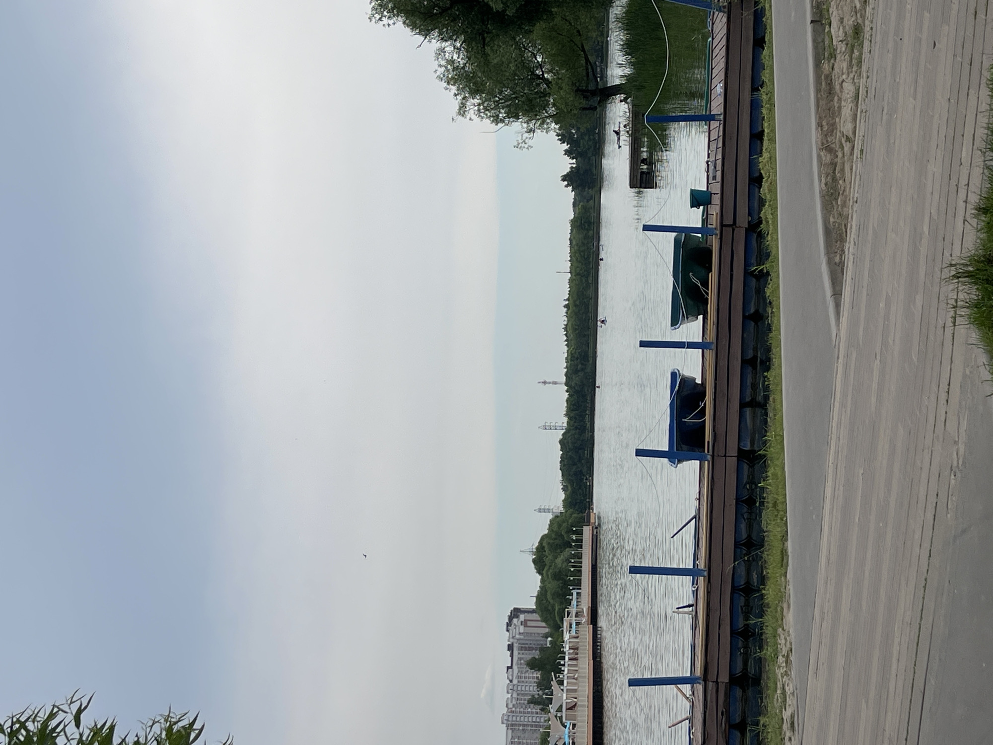 The image depicts a serene lake scene in a park, likely in Moscow, Russia. The sky is overcast, with a faint haze. In the foreground, a wooden boardwalk leads towards a floating dock. The dock is constructed from large, dark blue barrels supporting a wooden platform. Two blue posts with white rope are visible on the dock, along with two small boats. One boat is dark blue, and the other is green. A small blue bucket sits near the green boat.
Across the lake, a line of trees forms the horizon, with some buildings visible in the distance on the left, suggesting an urban park setting. Power lines and towers are also present in the background. Several individuals are scattered across the lake, some in kayaks and one possibly swimming. On the left side of the lake, a wide promenade area with white structures and seating is visible, possibly a beach or leisure area. A single bird is captured in flight in the upper part of the sky. The overall atmosphere is calm and suggests a recreational area within a city.