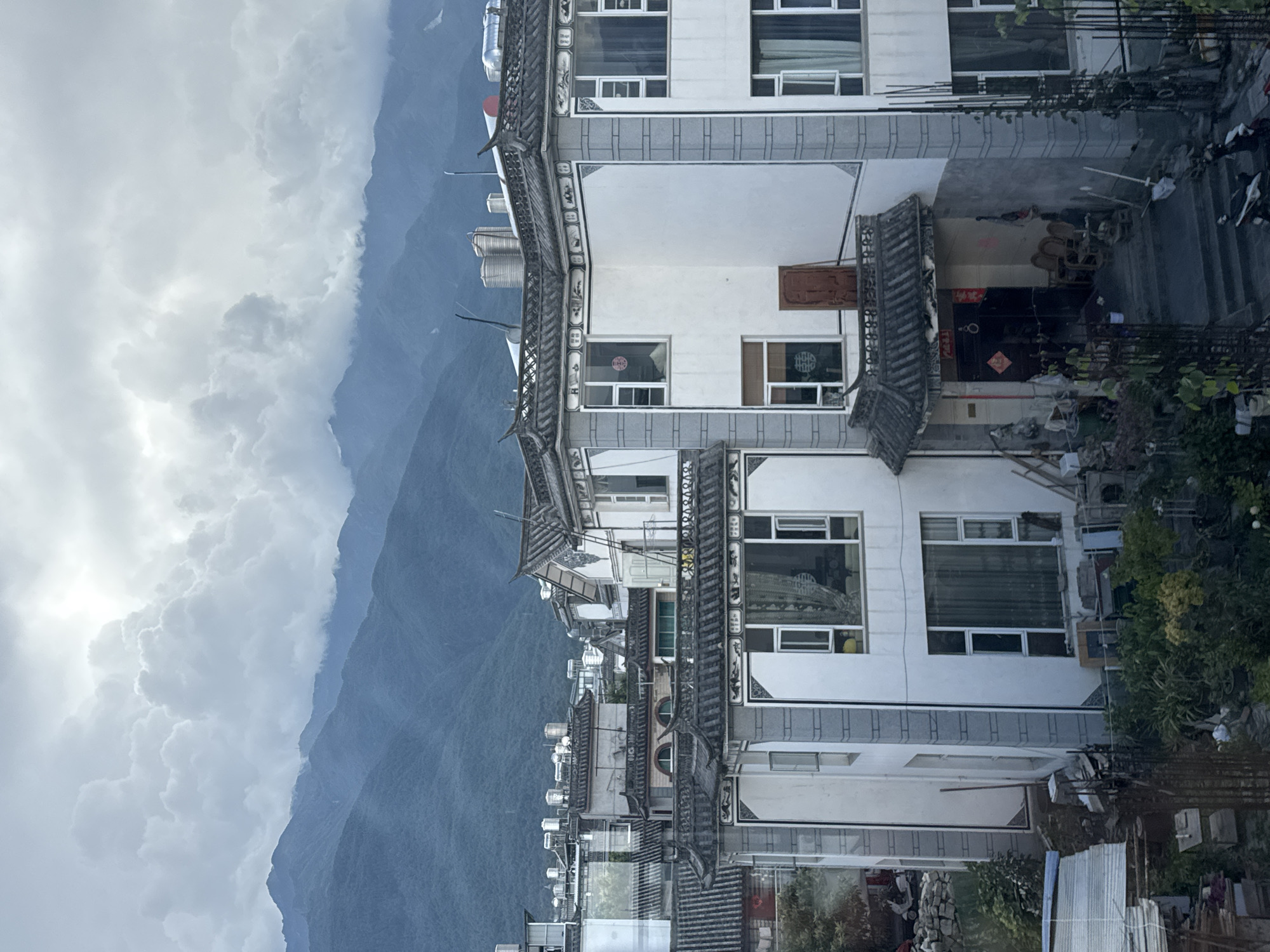 The image captures a view of residential buildings in Dali City, China, with a mountainous landscape in the background. The architecture of the buildings features traditional Chinese roof tiles and white facades with grey stone accents. Some buildings have multiple stories with various window designs. In the foreground, there is a cluttered area with construction materials, potted plants, and what appears to be a small scooter. The sky is overcast with dramatic clouds, and the mountains in the distance are shrouded in mist. The scene conveys a sense of everyday life in a semi-urban environment with a strong connection to the natural surroundings.