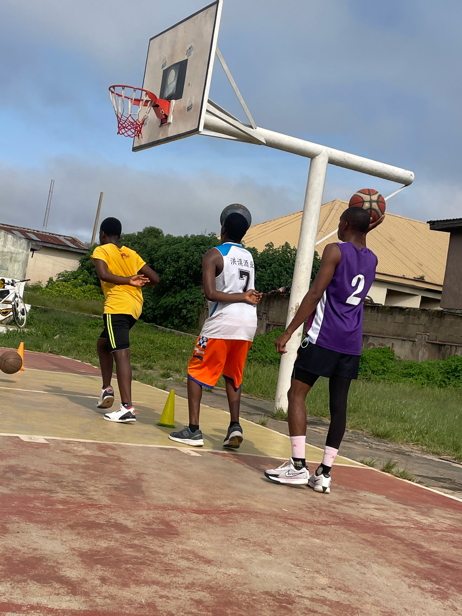 Three young males are on an outdoor basketball court in Gura Top, Nigeria. The court has a worn, reddish-brown surface with yellow markings. A basketball hoop with a white backboard and red net stands prominently. In the foreground, one individual in a purple jersey with the number '2' and black shorts holds a basketball above his head. To his left, another person in a white jersey with orange trim and orange shorts, also wearing a dark headband, is partially visible. The third person, to the far left, wears a bright yellow t-shirt and black shorts. Several yellow cones are scattered on the court, and a deflated basketball lies near the left edge. In the background, there are green trees, a low stone wall, and residential buildings with corrugated iron roofs, typical of the urban landscape in Gura Top. The sky is partly cloudy. A bicycle is parked on the left side of the image, near some structures.