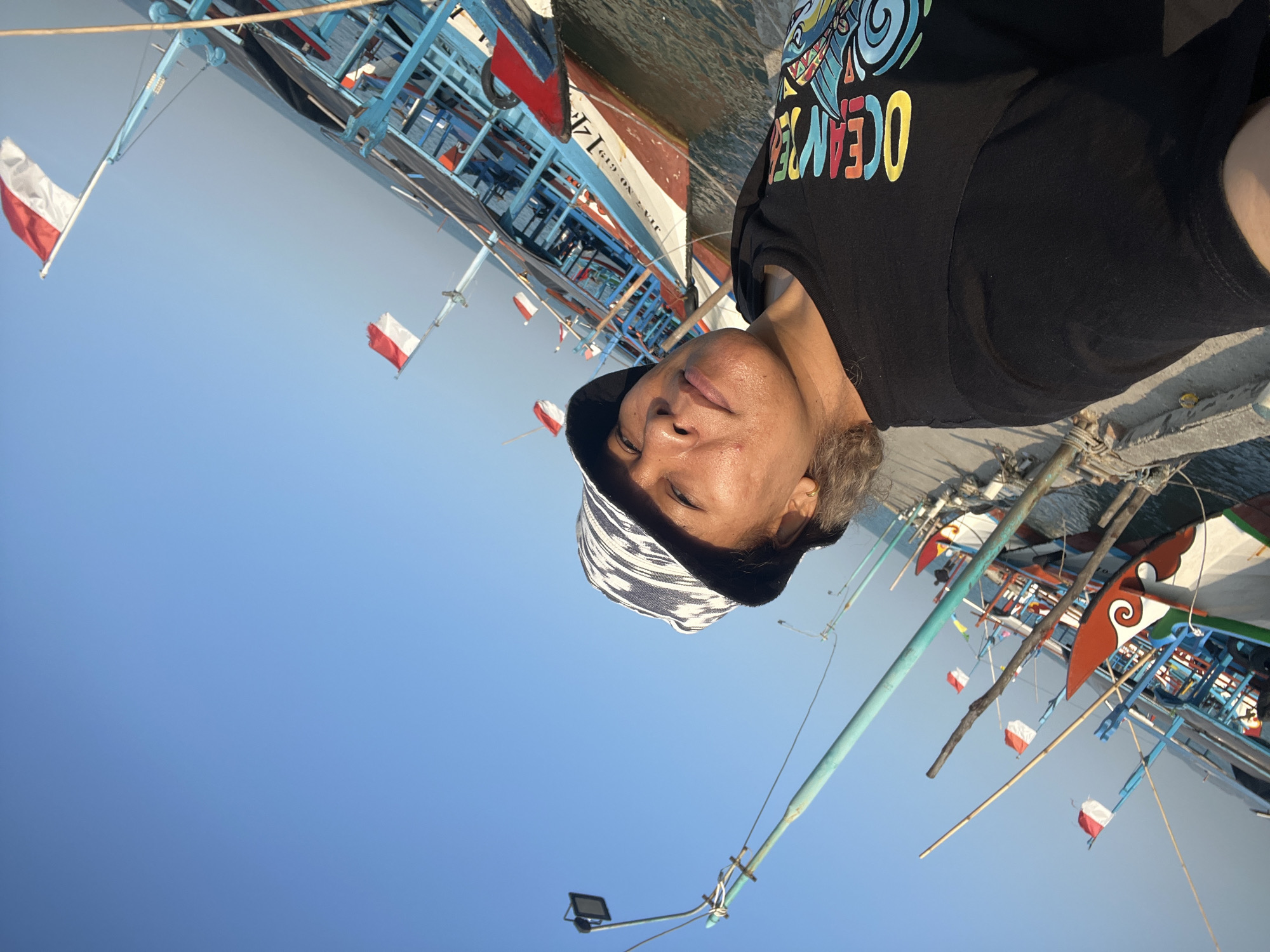 This image captures a selfie taken by a person wearing a black t-shirt with a colorful "OCEAN" graphic and a patterned bucket hat. The person is standing on a pier next to several Indonesian fishing boats, characterized by their blue and red hulls and the Indonesian flags displayed prominently. The sky is a clear, bright blue, suggesting a sunny day. In the background, other boats are docked, and the water is visible. Streetlights are also present along the pier. The scene is set in Jepara, Indonesia, a coastal city known for its fishing industry.