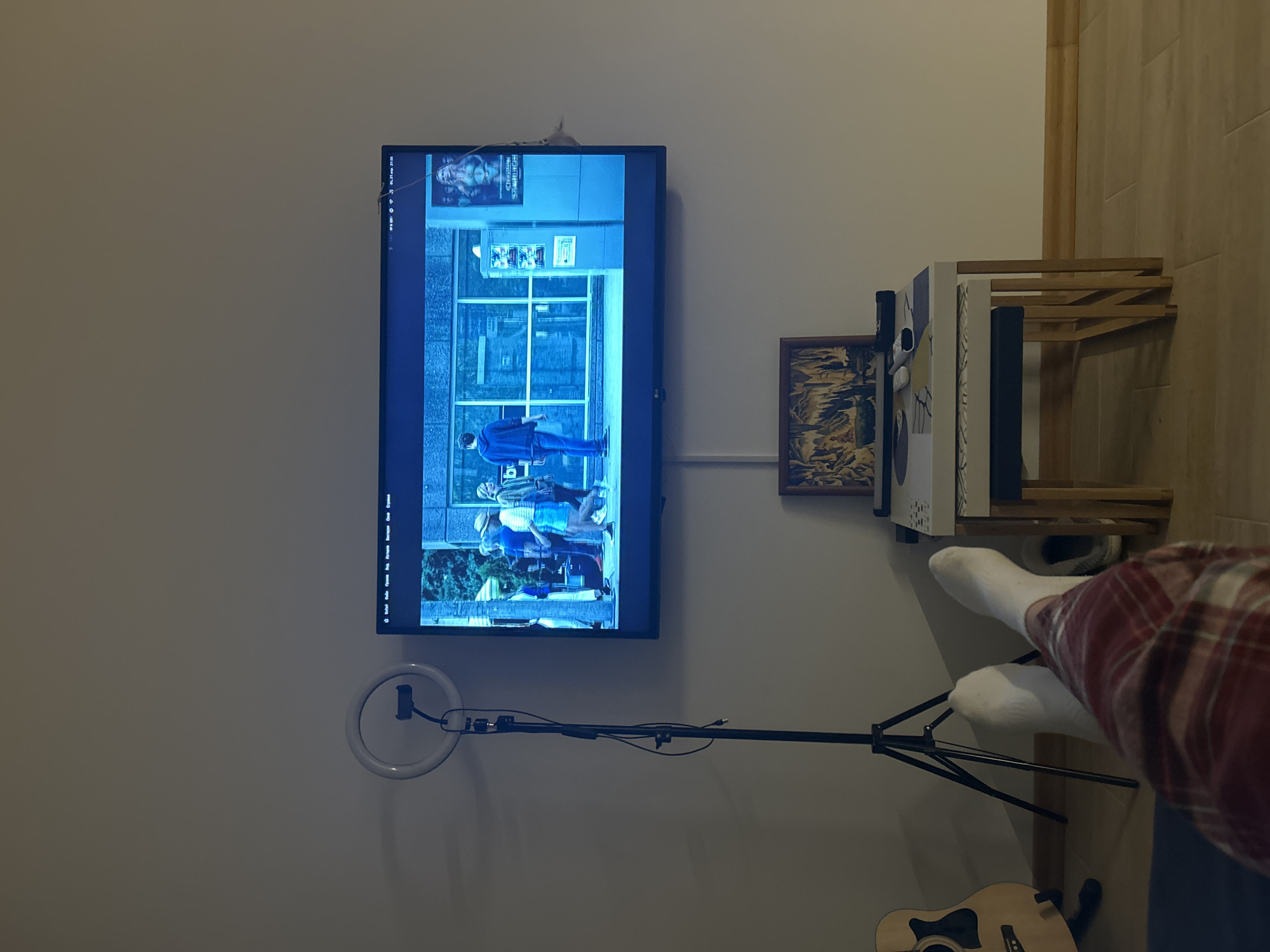 The image, captured from a tilted perspective and effectively rotated 90 degrees counter-clockwise, depicts a cozy indoor setting centered around a television screen. The wall-mounted television displays a scene with a strong blue tint, showing several people outdoors in front of a building with large glass windows. A poster on the screen, visible in the upper left corner of the displayed content, appears to read "CITIZEN" and "HAZARD". The television's interface, visible on the left side, includes menu options in a South Slavic language, such as "Gledaj" (Watch), "Info", "Podešavanja" (Settings), "Izvori" (Sources), "Snimanje" (Recording), "Pretraga" (Search), and "Čarobnjak" (Wizard).
Below the television, a white ring light with a smartphone holder is set up on a black tripod stand, positioned centrally. To the right of the television, a small, multi-tiered wooden shelf holds various items, and beneath it, a framed artwork or photograph with dark, possibly abstract or landscape imagery is hung.
In the foreground, partially visible in the bottom right, is the lower leg and foot of a person, covered in a red and white plaid fabric (likely a blanket or clothing) and a white sock, suggesting a relaxed viewing posture. An acoustic guitar with a light wood finish is also partially visible on the floor at the very bottom right. The room features light-colored walls and what appears to be a wooden floor along the right edge. The scene conveys a relaxed, domestic atmosphere, likely in the evening or night given the glow of the screen and the ring light.