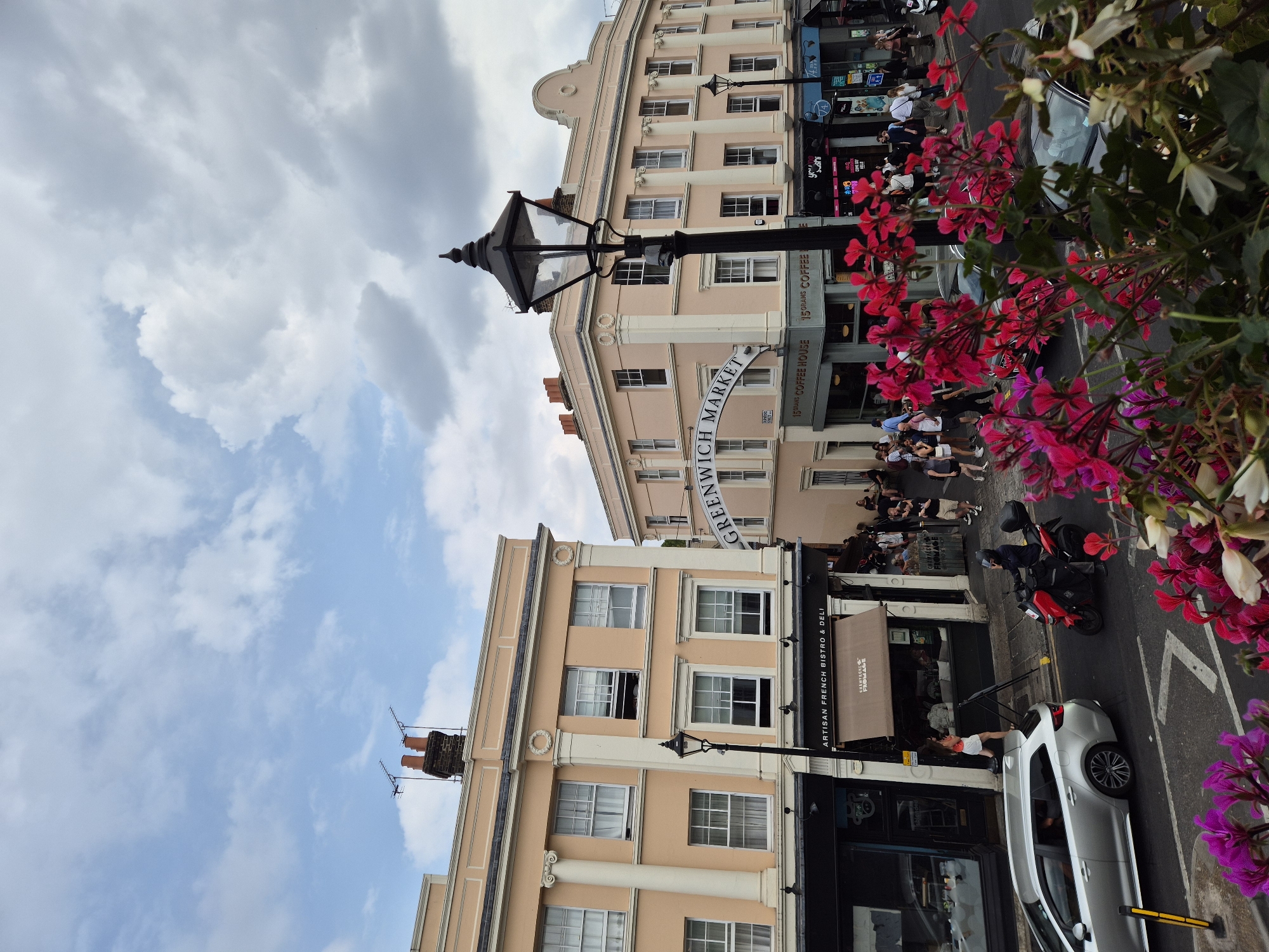 This is a high-angle, eye-level shot of Greenwich Market in London, United Kingdom, on a partly cloudy day. The scene depicts a busy street with pedestrians and vehicles.
In the foreground, vibrant pink and red flowers are in bloom, obscuring the lower part of the image. Behind the flowers, a silver station wagon is parked on the right side of the road. A red motorcycle is also visible, with a rider wearing a helmet.
The middle ground shows the facade of buildings lining the market. The building on the left is painted a pale peach color with white classical columns and decorative molding. It has multiple windows and a sign above a shop that reads "ARTISAN FRENCH BISTRO & DELI". A beige awning with the word "FIOMAGE" is extended over part of the storefront. The building on the right is also pale peach and features similar architectural elements, including columns and arched windows. A large, curved sign arching over the street reads "GREENWICH MARKET". Below this sign, several shops are visible, including "15 GRAMS COFFEE HOUSE".
The background consists of a sky with a mix of blue patches and fluffy grey clouds. The rooftops of the buildings show chimneys and antennae. A black streetlamp with a decorative lantern stands prominently in the mid-ground.
People are seen walking along the market street, some are standing and chatting, and others are seated outside a cafe. The overall impression is one of a lively urban marketplace.