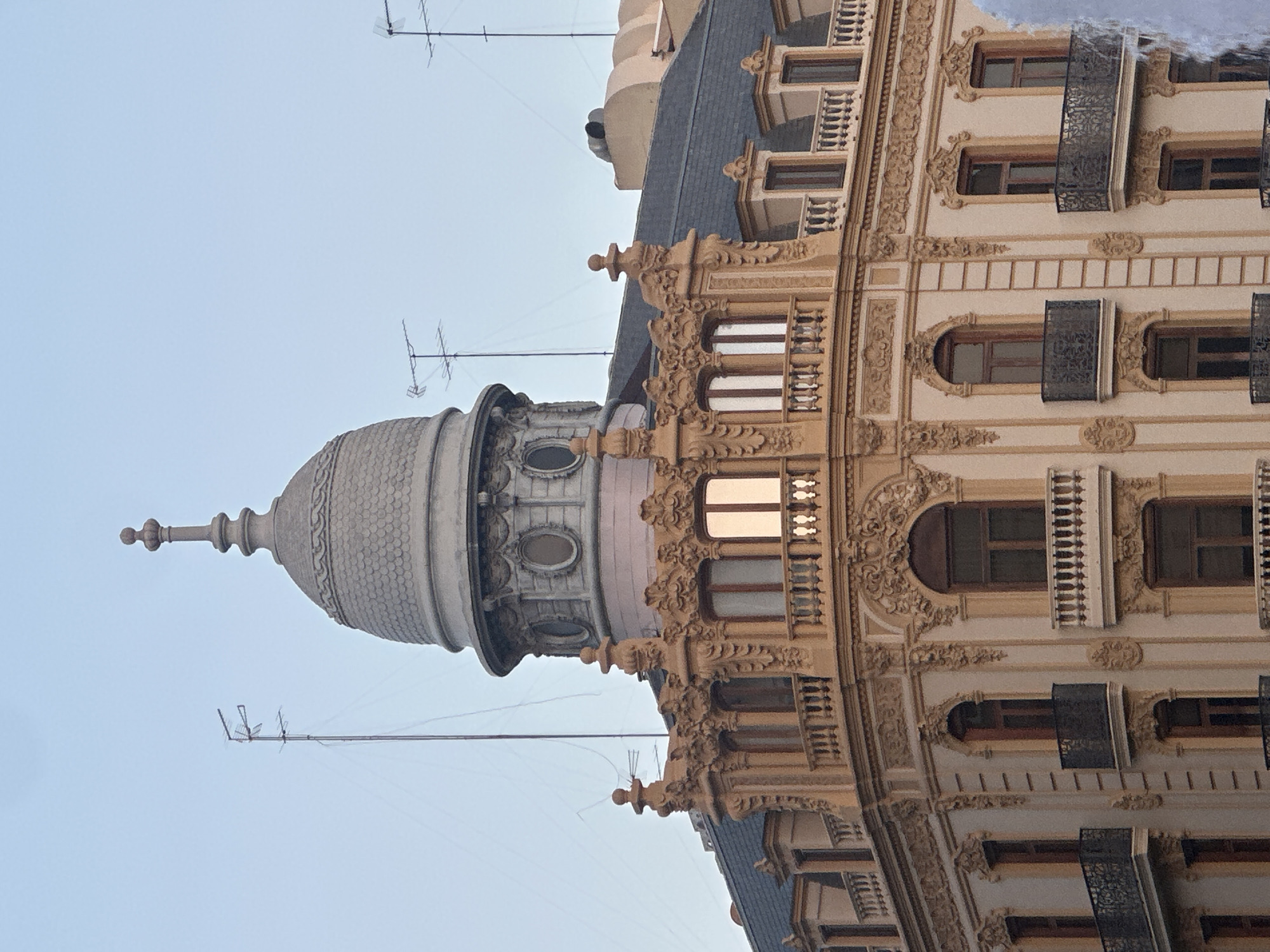 The image shows the upper facade of a building in Valencia, Spain, with an ornate architectural style. The building features a domed turret topped with a finial, several levels of balconies with decorative railings, and numerous windows framed by elaborate moldings. The facade is painted in a light beige or cream color, with contrasting darker trim around windows and balconies. The roof is covered in dark tiles. Several television antennas are visible on the rooftop. The sky is a clear, pale blue. The architecture suggests a historic building, possibly from the late 19th or early 20th century.
