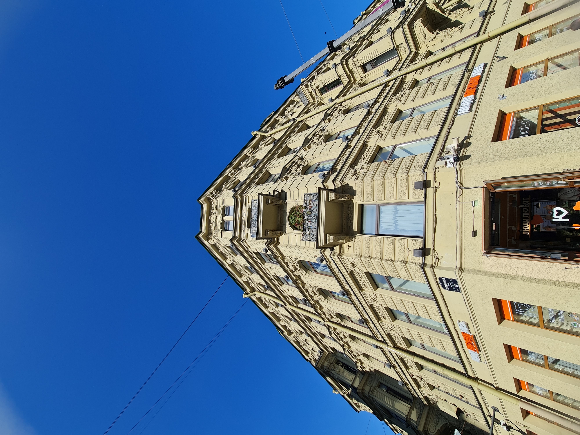 A low-angle, wide shot captures the facade of a light yellow, multi-story building under a clear, vibrant blue sky in Saint Petersburg, Russia. The building features ornate architectural details, including intricate carvings around the windows and cornices, as well as decorative balconies. Several windows are visible, some with curtains. Yellow drainpipes run vertically along the exterior.
At street level, an open doorway reveals a glimpse of an interior with signage. Orange-colored window frames are prominent on the lower floors, and some windows display text, such as "ВСЁ" which translates to "everything," and an image of a heart. A sign on the building reads "КАРАВАННАЯ ул. № 28," indicating the street name and building number.
Electrical wires stretch across the sky in the upper left portion of the image. A tall, grey pole with a dark top extends upwards from the right side of the building, obscuring part of the sky and the building's upper right section. The lighting suggests it is daytime, with sunlight illuminating the facade. No people are clearly visible in the scene.