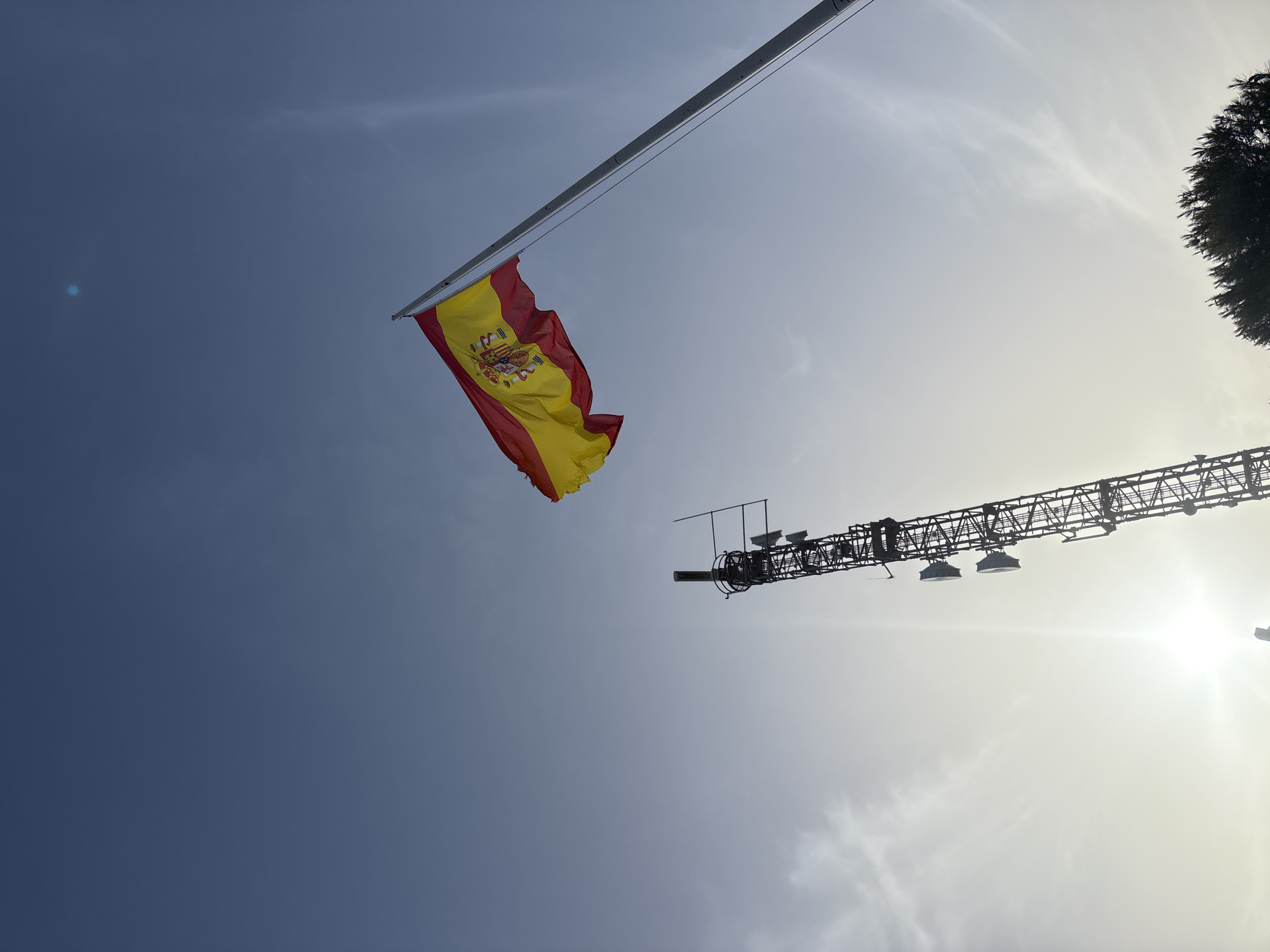 The image shows a Spanish flag waving on a flagpole against a clear blue sky with wispy clouds. To the left of the flag is a tall, metallic telecommunications tower with multiple antennae attached. The sun is visible as a bright orb in the lower left portion of the image, casting a bright glare. The scene appears to be outdoors, with a treeline visible at the bottom edge of the frame. The location is identified as Orihuela, Spain.