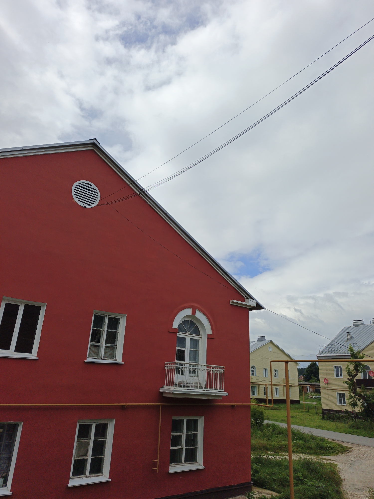 This is a medium-shot, eye-level, full-color photograph taken on an overcast day in Medvedyovka, Russia. The foreground is dominated by a bright red building with a steeply pitched roof. Several white-framed windows are visible on the building's facade, some appearing slightly askew. A circular ventilation grate is present high on the wall, near the roofline. A small white balcony with a railing protrudes from the upper story. A yellow pipe runs horizontally across the red building's exterior. In the background, two pale yellow buildings with grey roofs are visible. A paved road curves through the scene, bordered by patchy green grass and some trees. Overhead, three dark power lines stretch diagonally across the cloudy sky. No people or animals are present in the image.