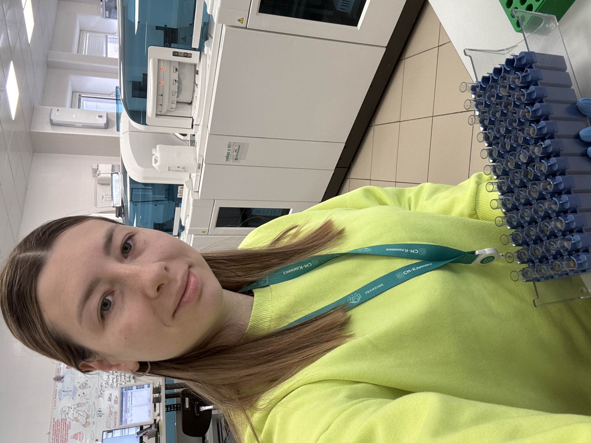 A young woman is captured in a selfie within a brightly lit medical laboratory, likely in Moscow, Russia, given the Cyrillic text. She wears a bright lime-green sweatshirt and a green lanyard. The lanyard prominently displays "СМ-Клиника" (SM-Klinika), a known medical clinic network in Russia, and "УВАЖЕНИЕ" (Respect).
Behind her, the setting is dominated by large, white and blue diagnostic equipment, indicative of a clinical laboratory. One prominent piece of machinery clearly shows the model "cobas e 801," a high-volume immunoassay analyzer manufactured by Roche Diagnostics. To the right of the woman, a clear rack holds numerous blue-capped test tubes or vials, suggesting active sample processing or storage.
In the background, to the left, a whiteboard is visible with various handwritten notes and simple drawings. Visible text on the whiteboard includes "С днем рема" (Happy Rem Day), "4/2 Контроли" (4/2 Controls), and "PCR" (Polymerase Chain Reaction), along with other technical notations and diagrams. The woman has a neutral to slightly smiling expression, and the scene appears to take place during the daytime, with ample natural light from windows. The tiled floor and clean, functional environment reinforce the laboratory setting.