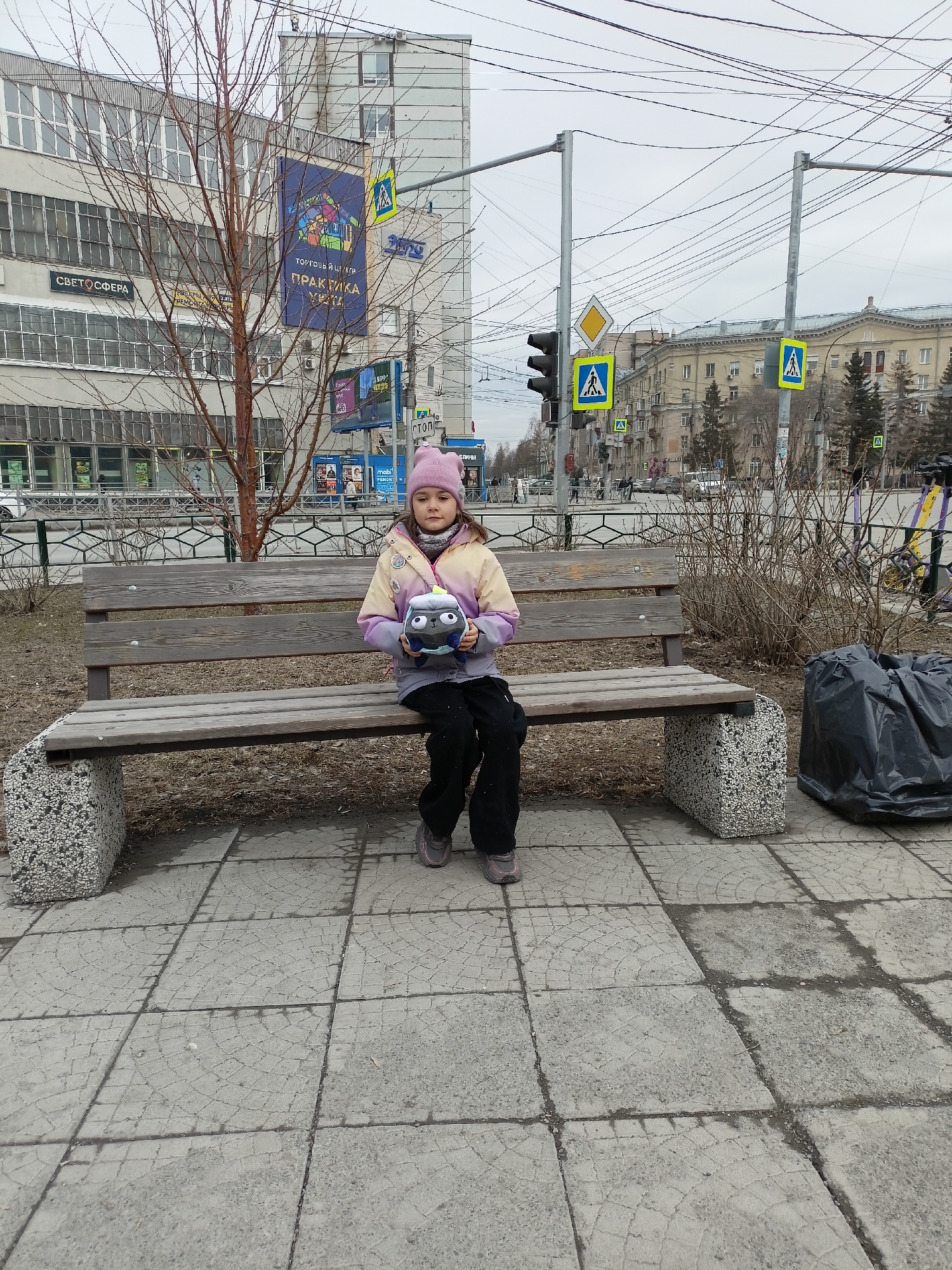 A young girl wearing a pink hat, a multi-colored jacket, and dark pants is sitting on a wooden bench in a city environment. She is holding a plush toy. Behind the bench, there is a bare tree and some dry bushes. To the right of the bench, there is a black plastic garbage bag. The ground is paved with gray concrete tiles. In the background, there are buildings, pedestrian crossing signs, traffic lights, and overhead power lines, typical of an urban setting. The building directly behind the tree has a sign in Russian that includes the words "ТОРГОВЫЙ ЦЕНТР" (shopping center) and "ПРАКТИКА" (practice). Another sign on the same building reads "СВЕТОСФЕРА" (svetosphera). Considering the building architecture, street signs, and language on the signs, the location is likely in Novosibirsk, Russia.